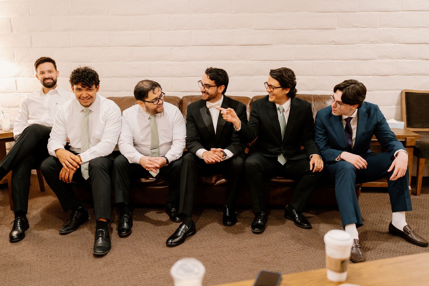 Six groomsmen in suits laughing and relaxing together on leather couch before wedding ceremony.