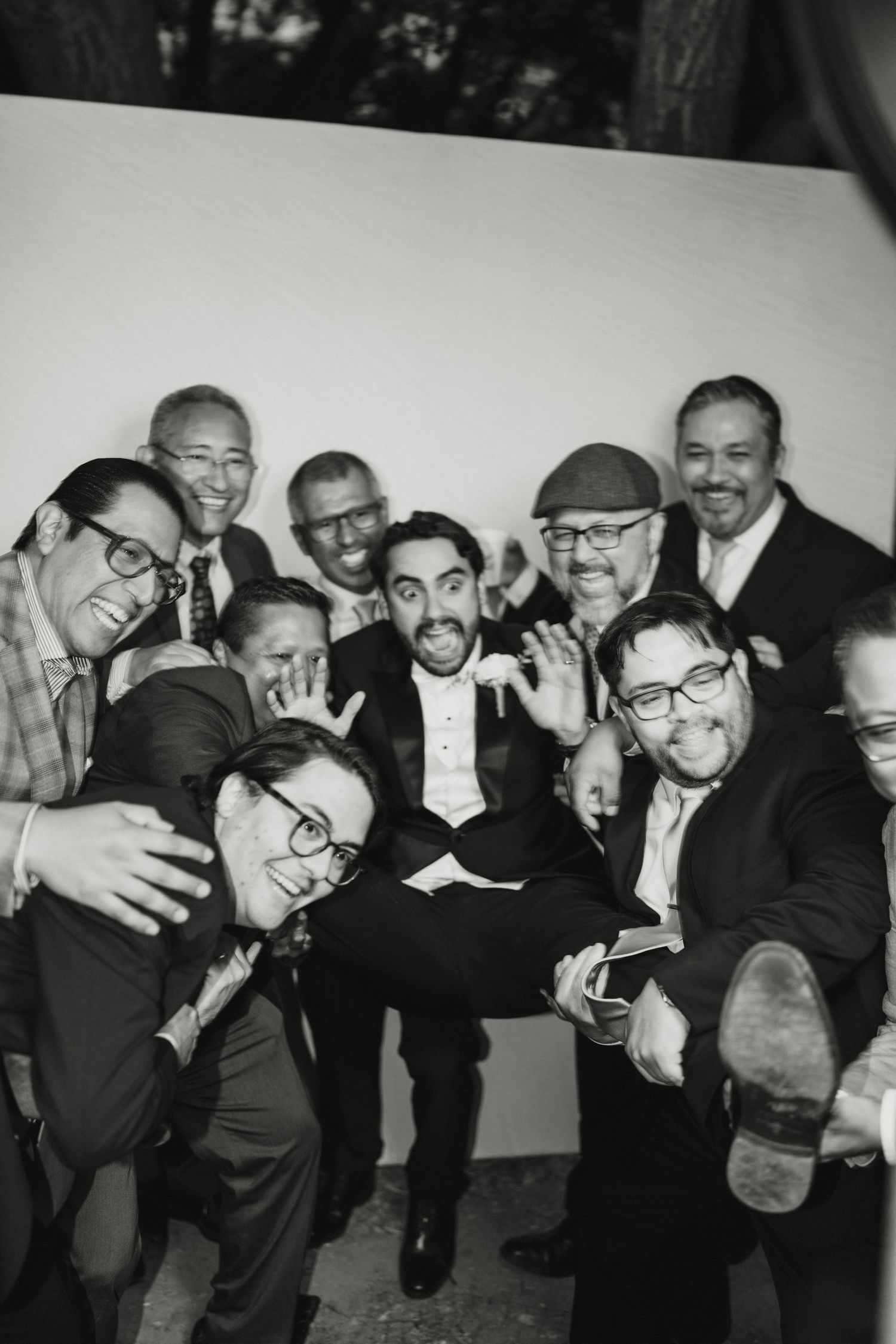 Black and white photo of groom surrounded by laughing groomsmen in a fun candid wedding photo booth moment.