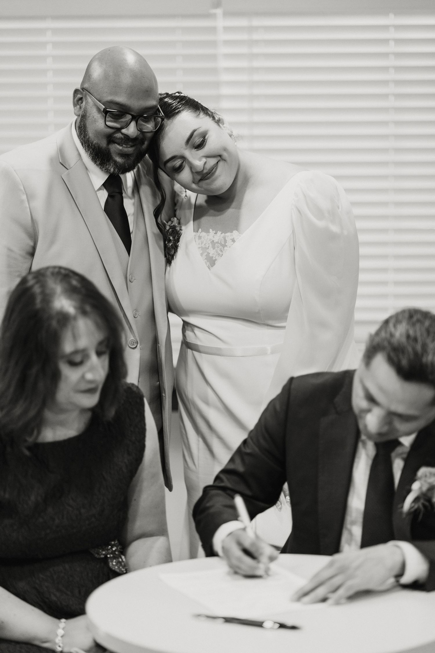 Black and white photo of newlyweds embracing while witnesses sign marriage certificate.