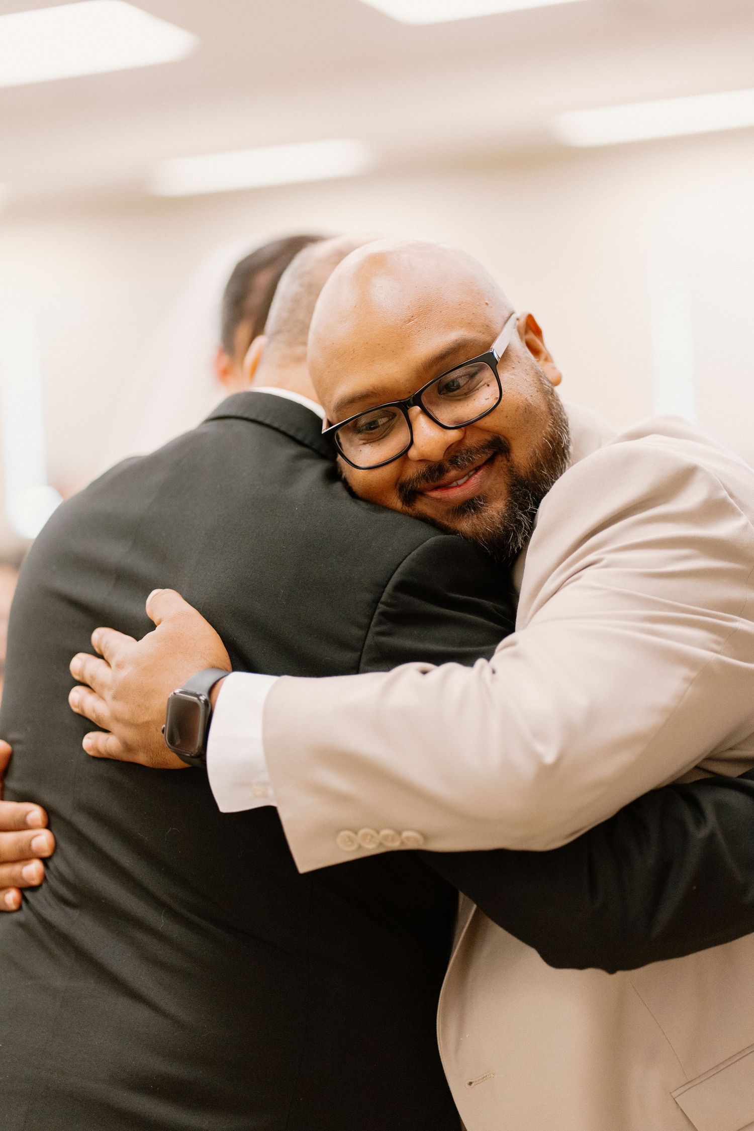 Smiling groom in beige suit and glasses shares a warm hug with a guest at his wedding ceremony.