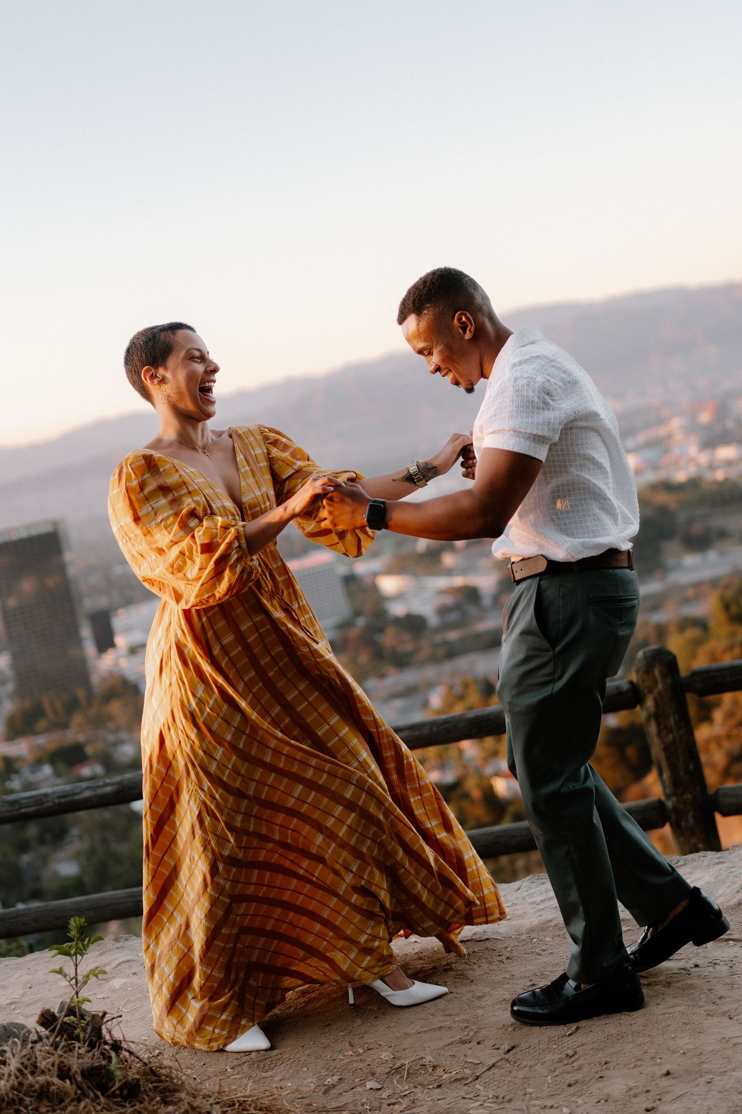 Couple dancing joyfully at sunset on a hilltop, woman in flowing orange dress, city skyline in background.