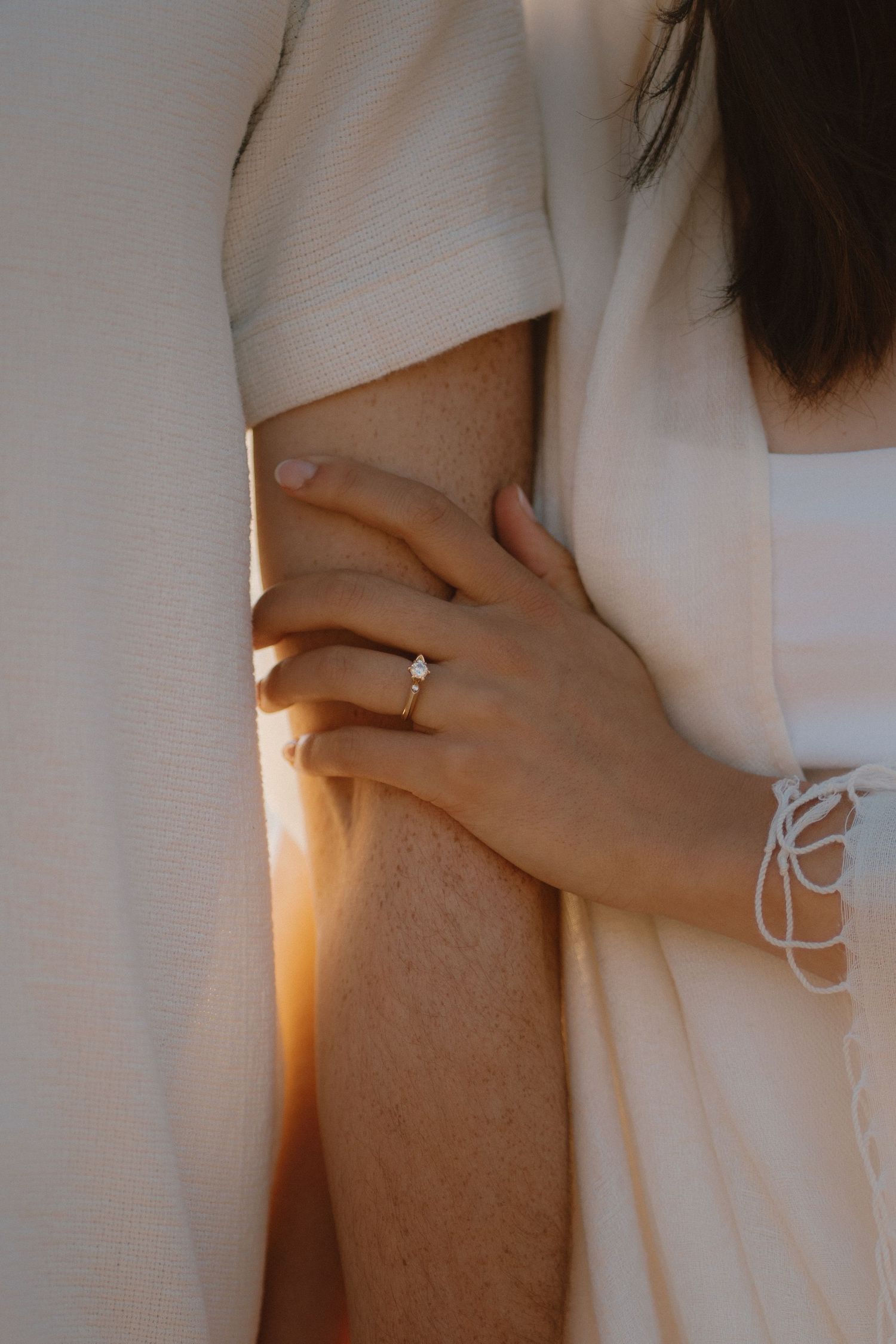 Close-up of a woman wearing an engagement ring gently holding a mans arm in warm golden light.