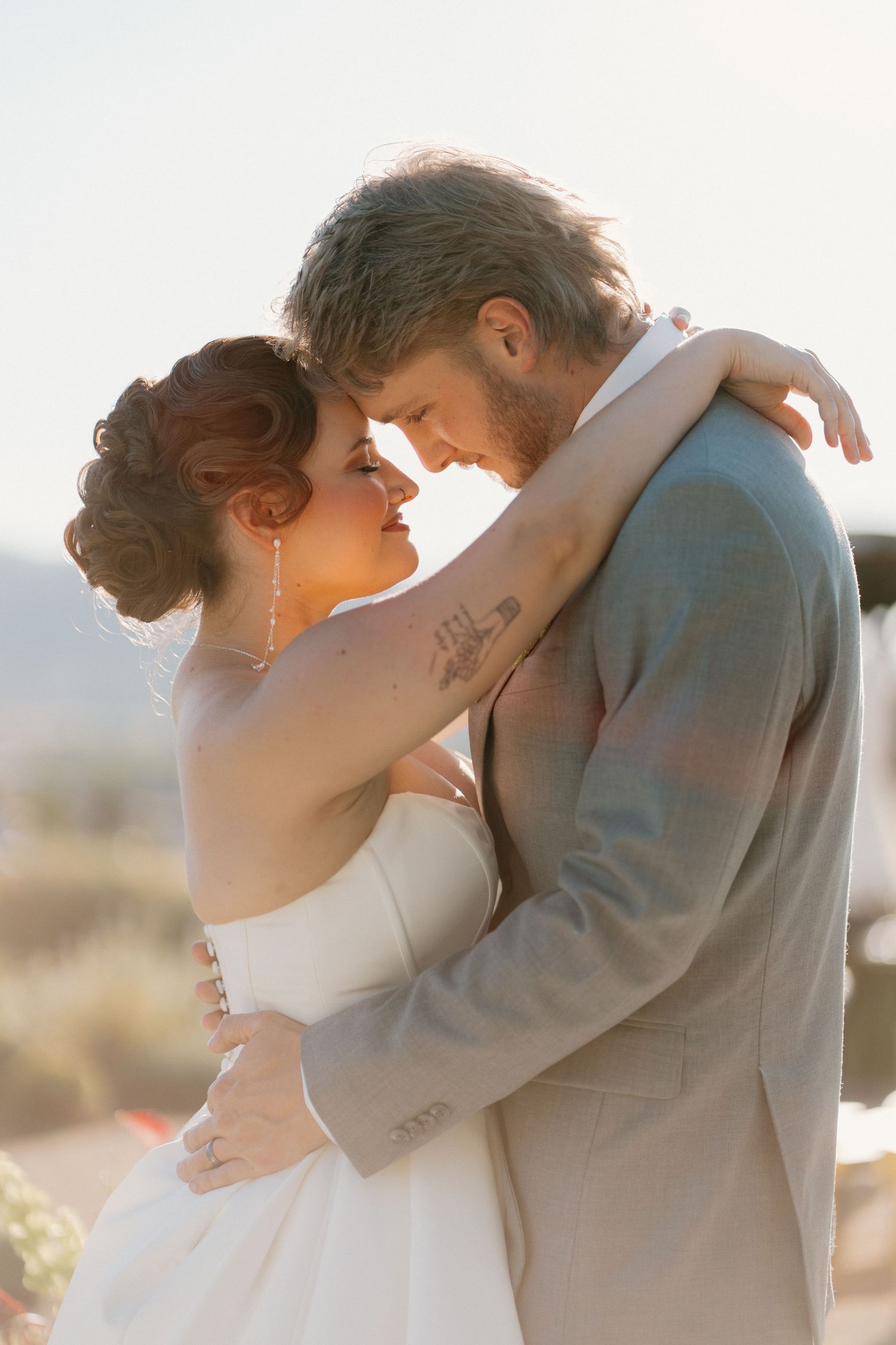 Bride and groom embracing forehead-to-forehead in golden backlight during outdoor wedding ceremony.