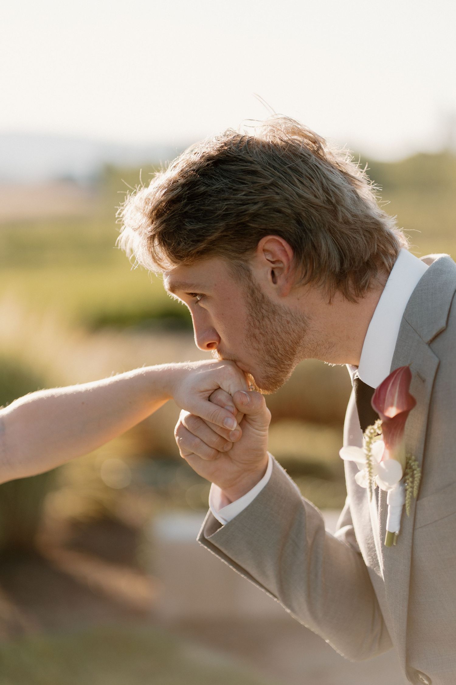 Groom in a tan suit gallantly kisses the hand of his bride during their outdoor wedding ceremony.