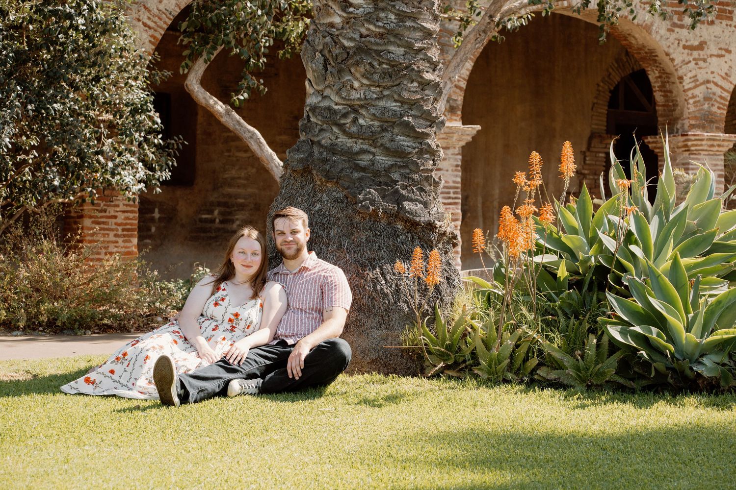 Couple sitting on grass in front of brick arches and lush gardens at Mission San Juan Capistrano.