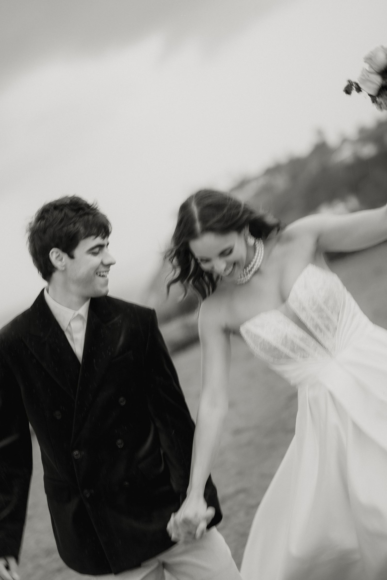 Black and white photo of laughing newlyweds holding hands outdoors, bride in flowing white gown and groom in dark velvet blazer.
