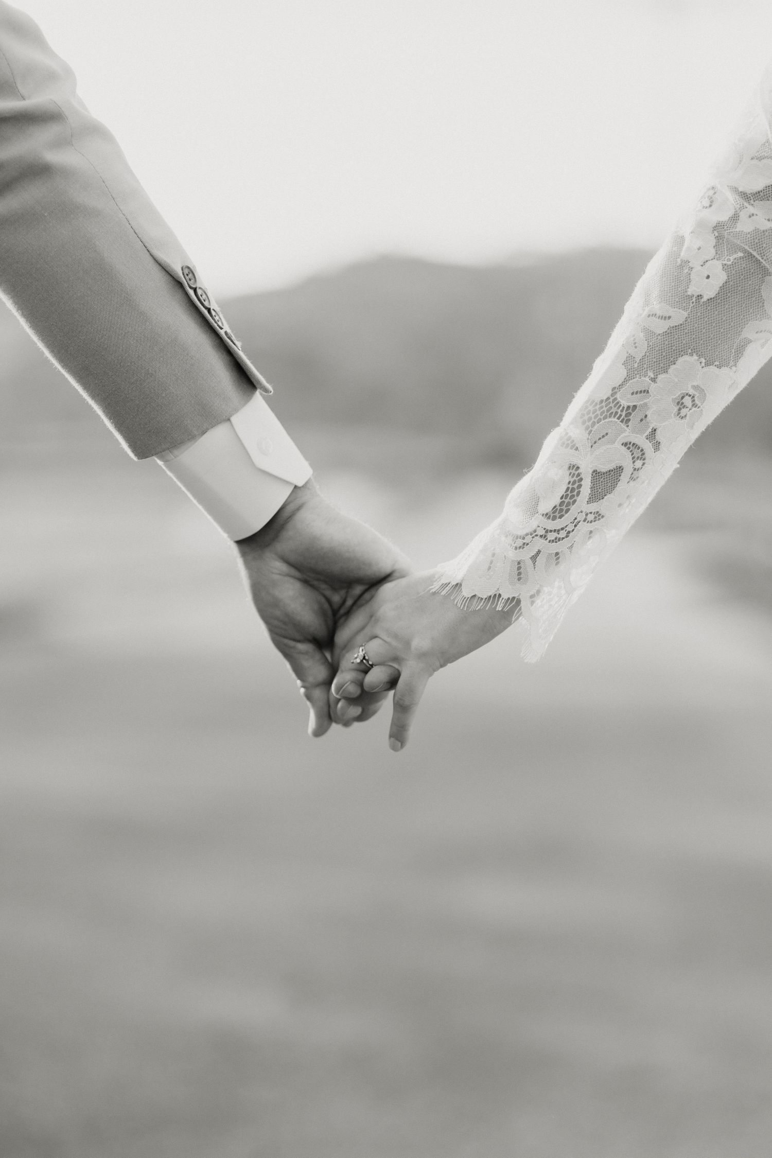 Black and white close-up of bride and groom holding hands, showcasing lace wedding dress sleeve.
