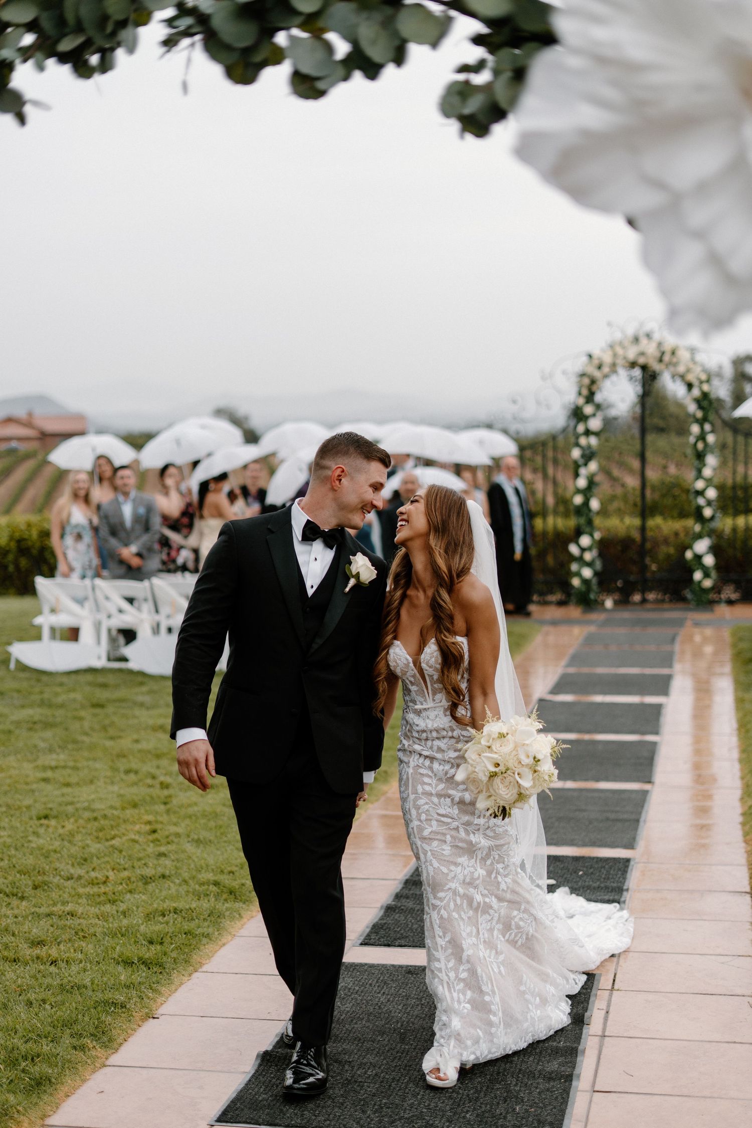 Newlywed couple walking down the aisle after their outdoor wedding ceremony as guests hold white umbrellas.