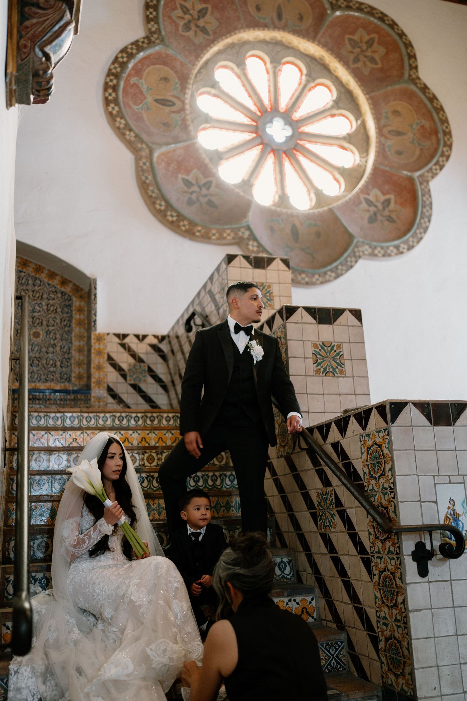 Bride and groom on decorative tiled staircase beneath stunning rose window in historic venue.