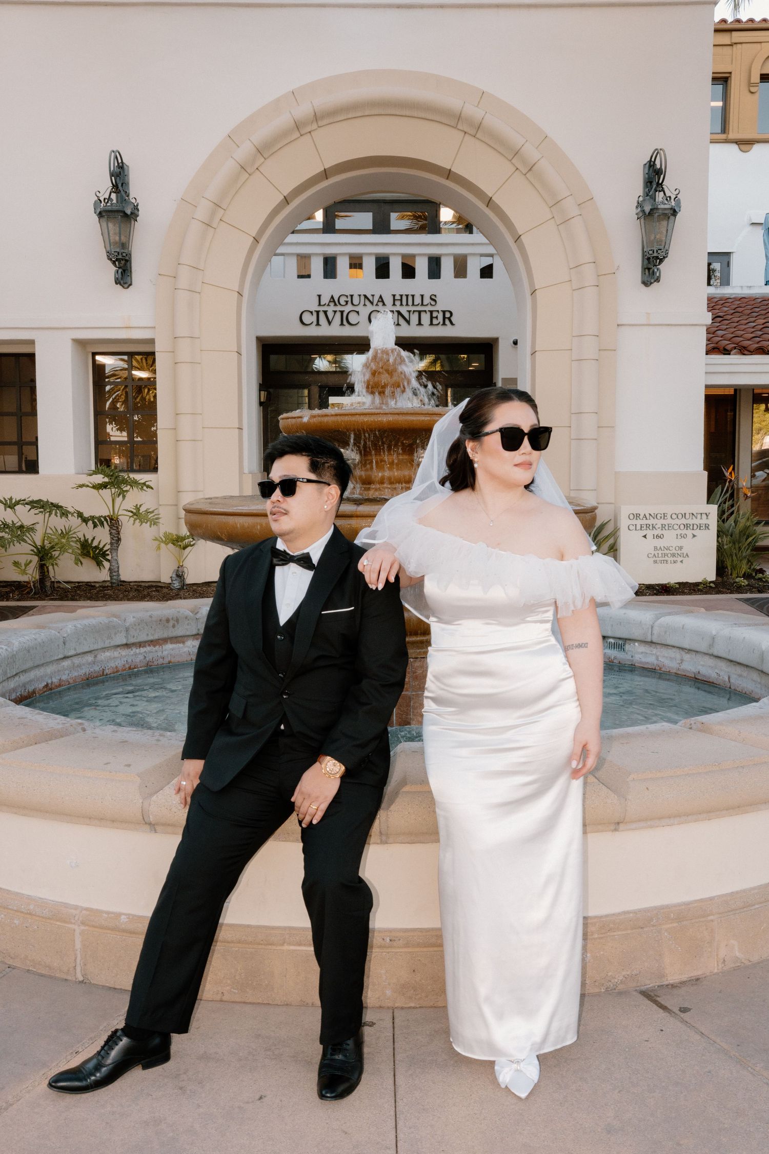 Stylish newlyweds posing with sunglasses at Laguna Hills Civic Center fountain.