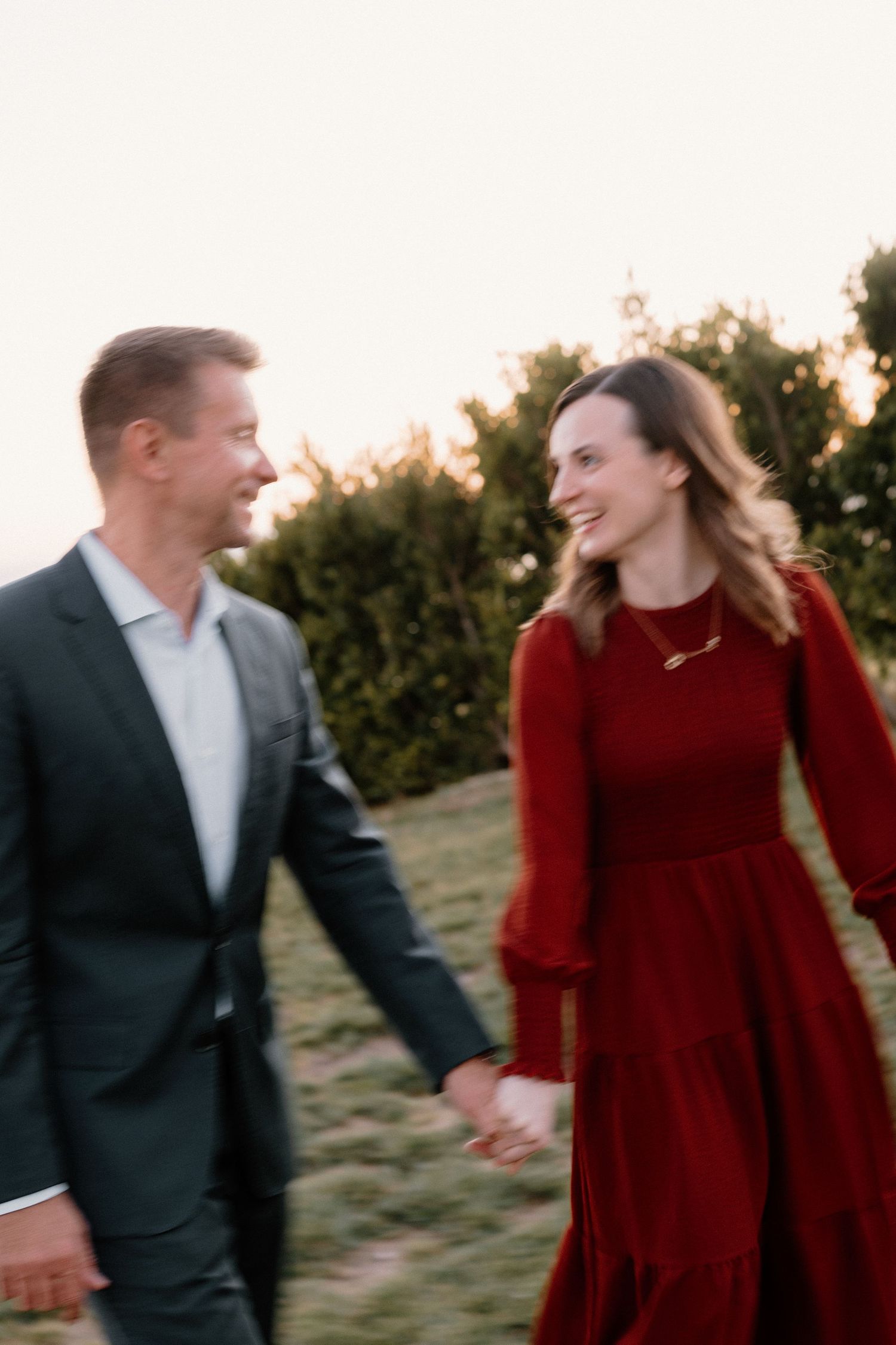 Happy couple holding hands and walking through a garden at golden hour, woman in red dress.