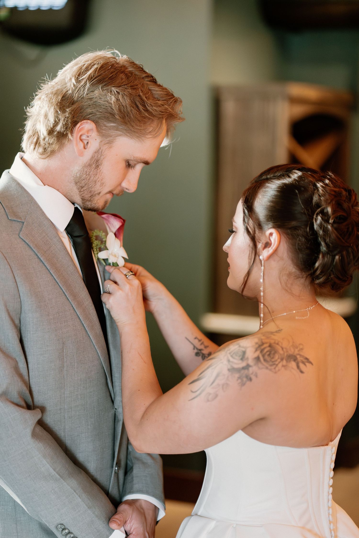 Bride with tattoos pins a red calla lily boutonniere on groom in gray suit before wedding ceremony.
