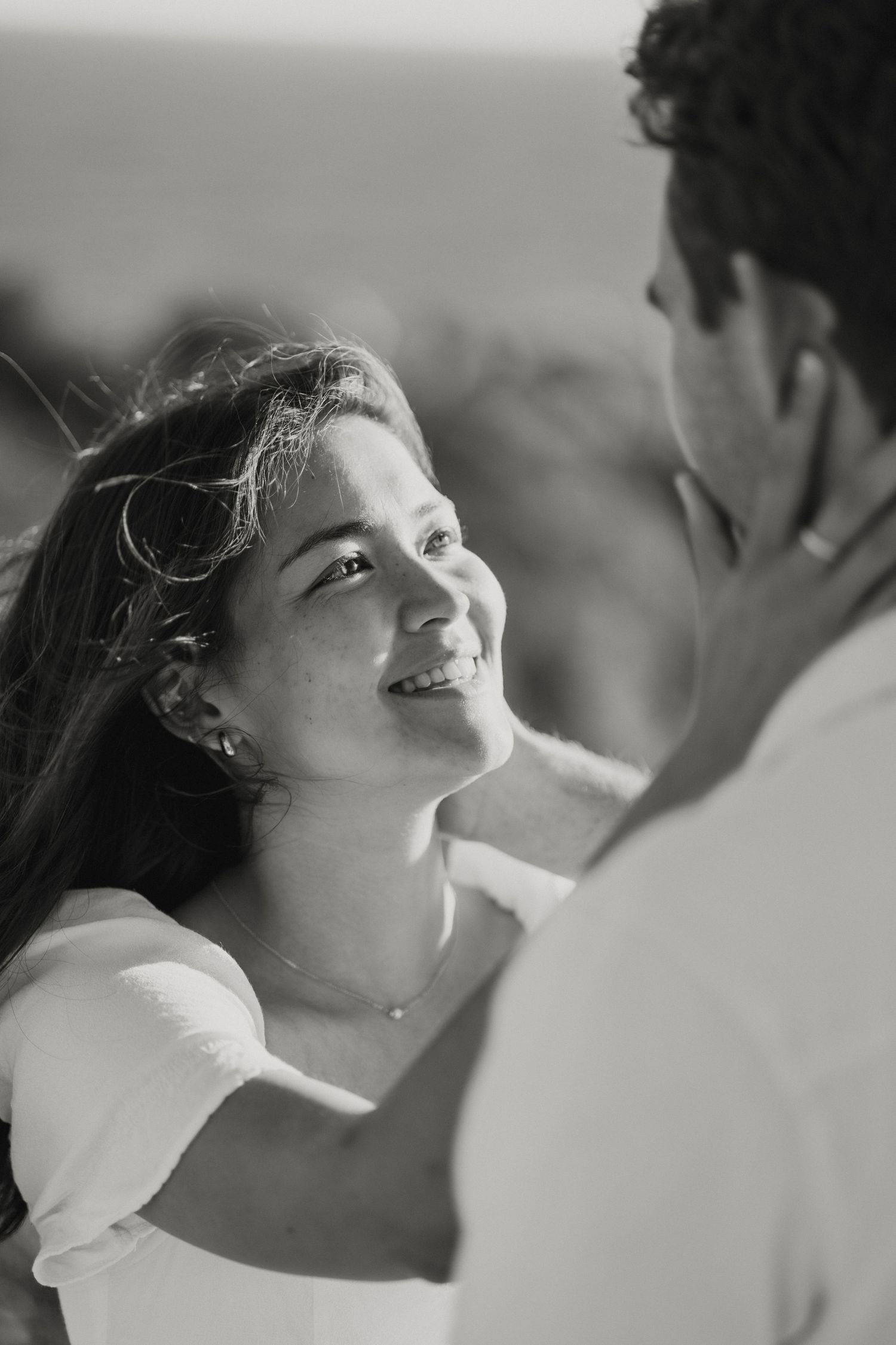Black and white portrait of a smiling woman gazing lovingly at her partner on a breezy beach.