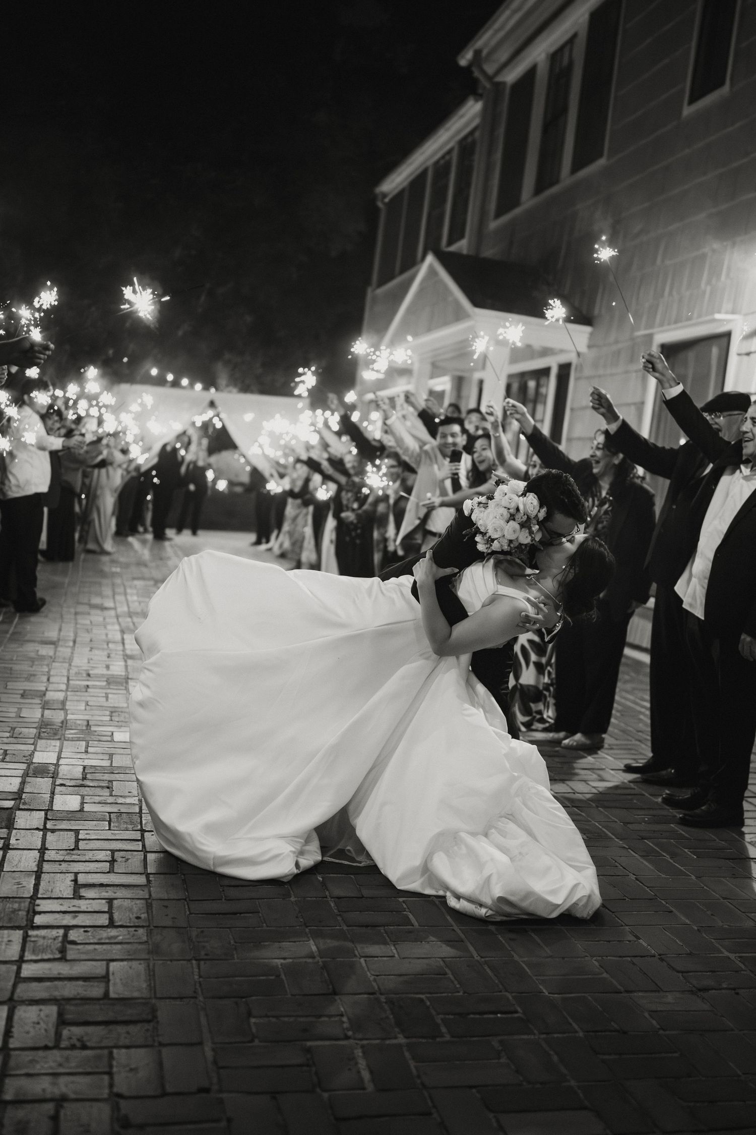 Black and white photo of bride and groom sharing dramatic dip kiss during sparkler wedding sendoff.
