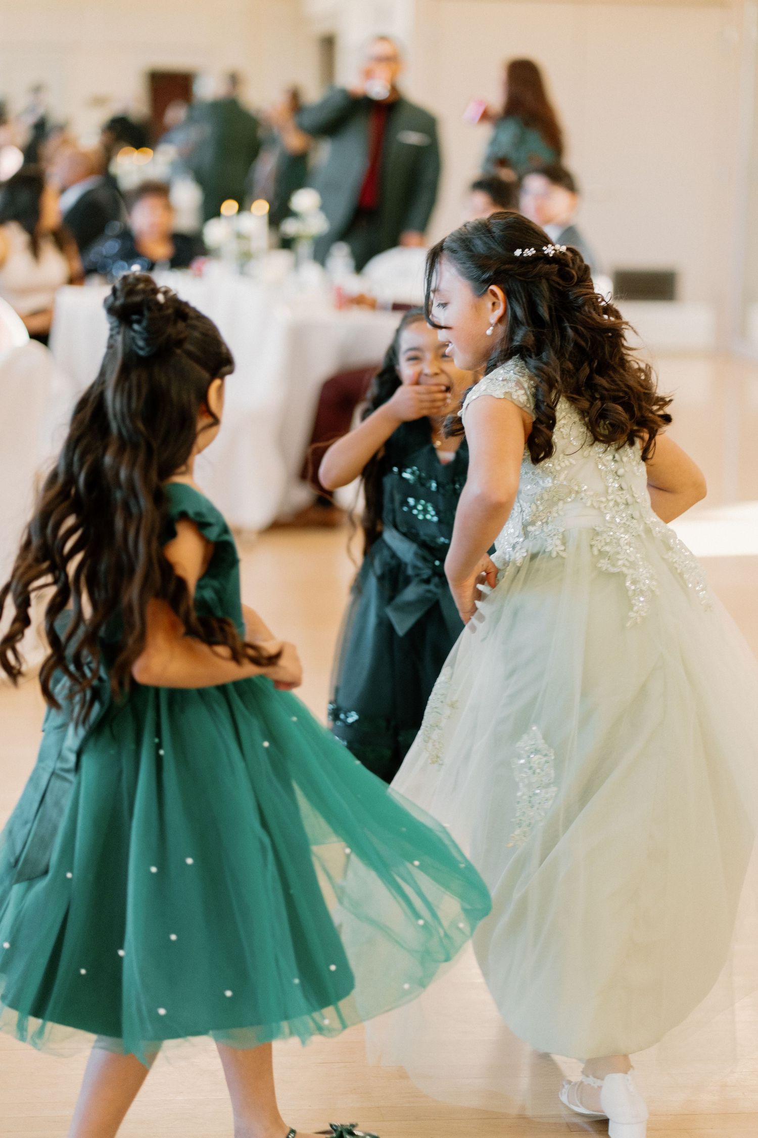 Young flower girls in green dresses dance joyfully at an elegant wedding reception.