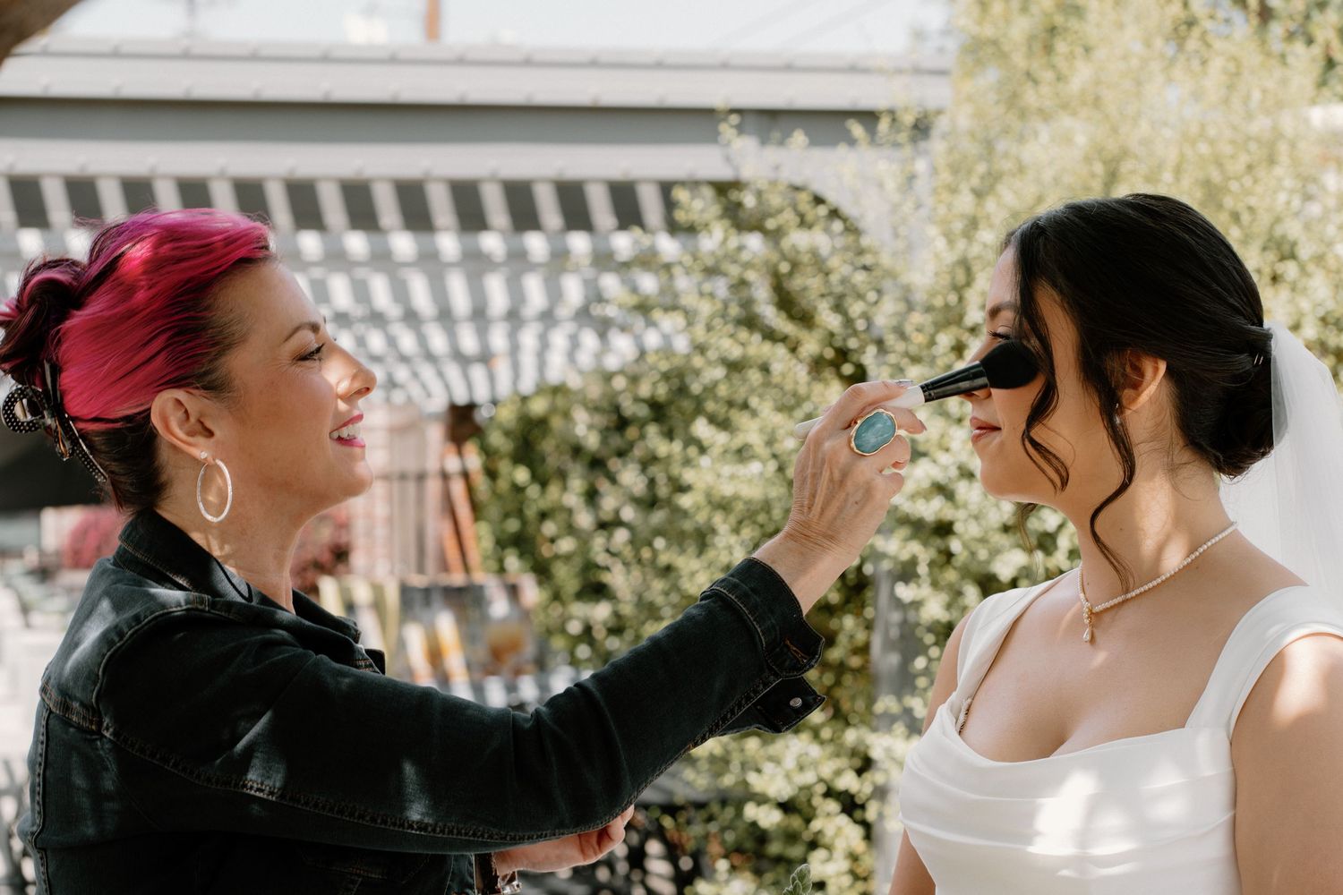 Makeup artist with pink hair applying makeup to a bride in a white dress outdoors before her wedding.