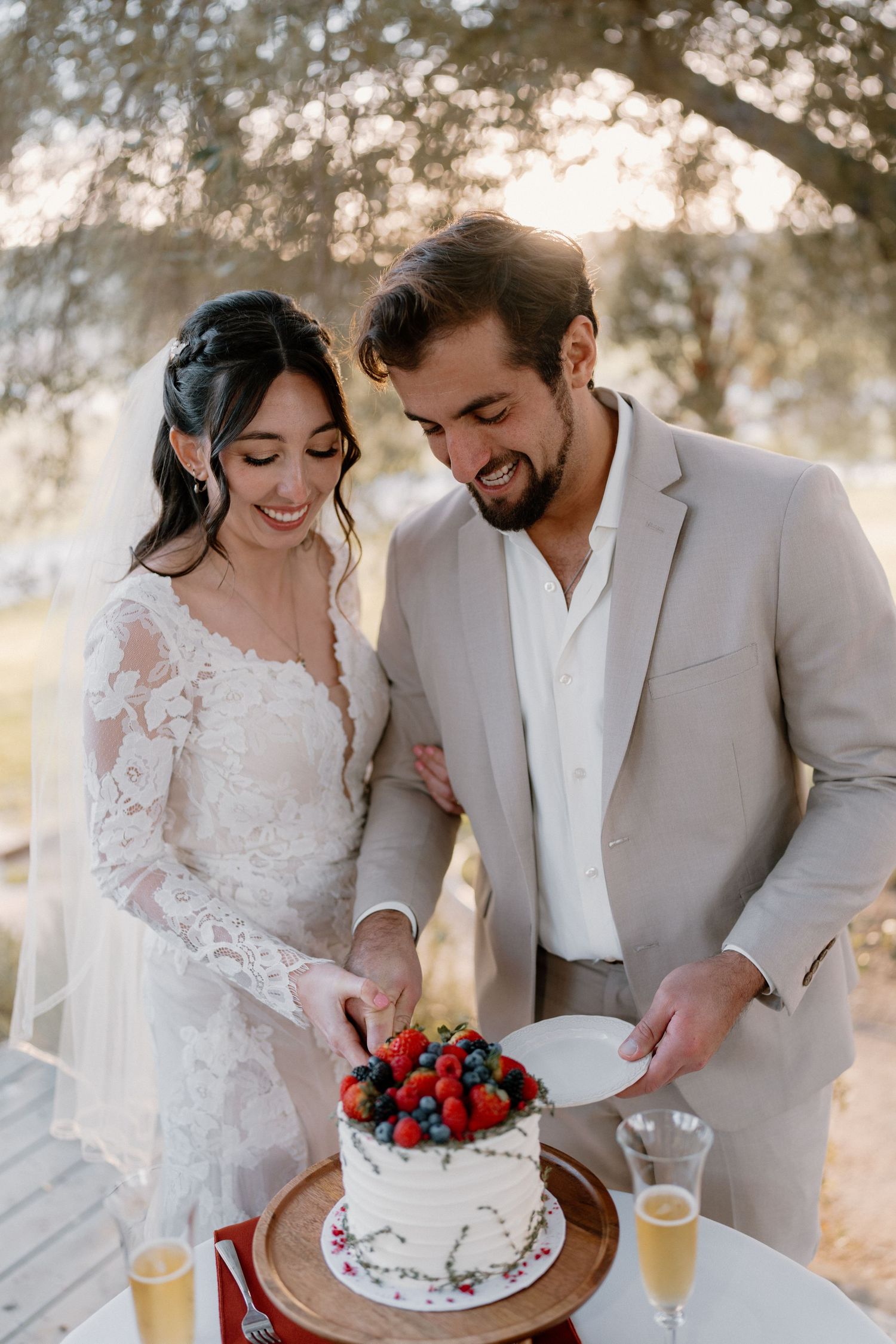 Smiling bride and groom in beige suit cutting white berry-topped wedding cake at outdoor reception.