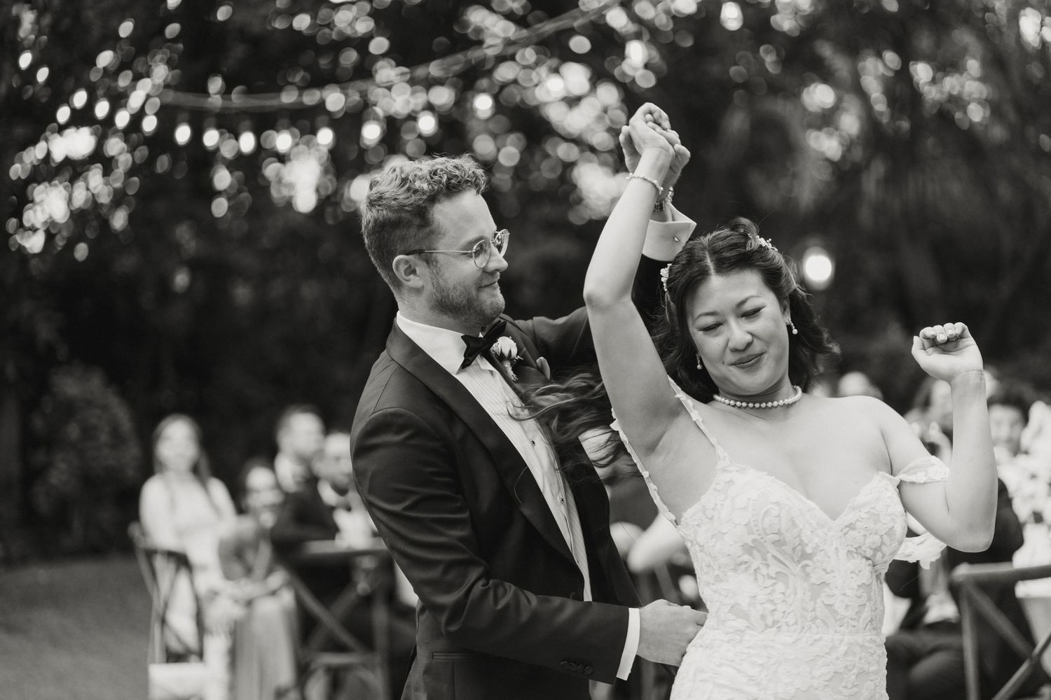 Black and white photo of groom spinning bride joyfully during outdoor wedding ceremony with string lights.