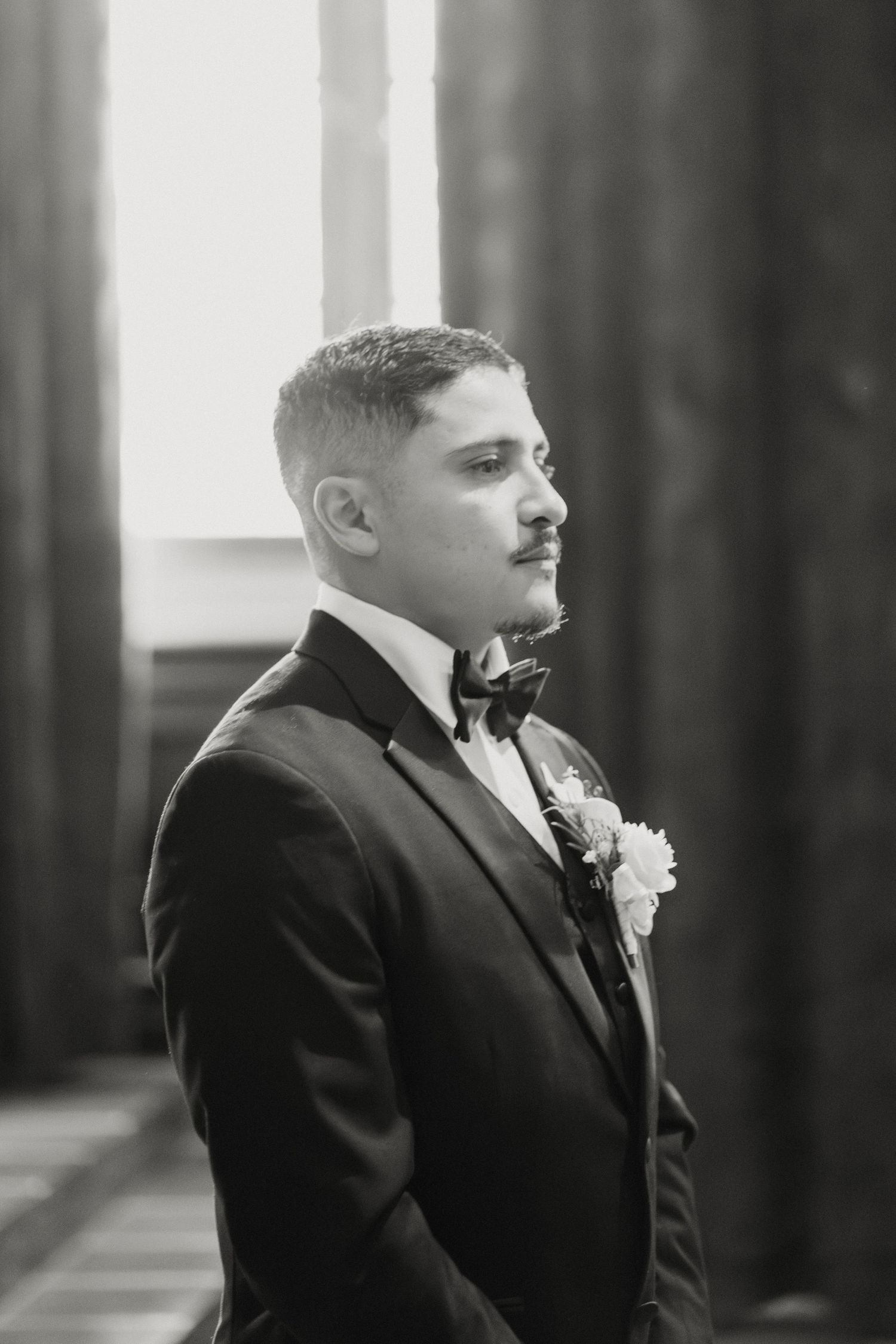 Groom in black tuxedo with bow tie and boutonniere gazing thoughtfully in black and white wedding photo.