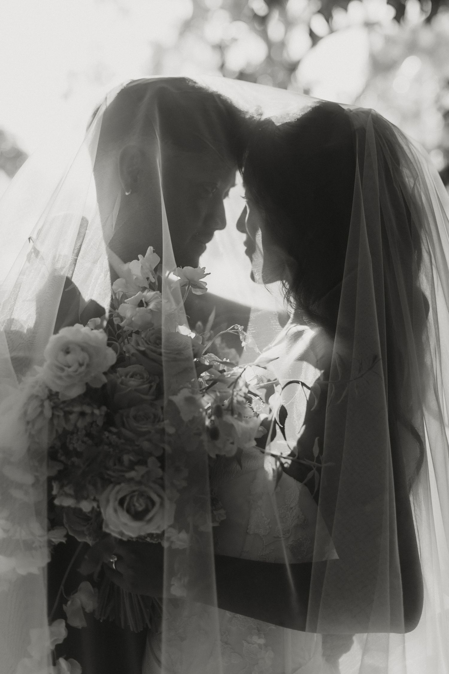Romantic black and white portrait of bride and groom hidden beneath wedding veil holding floral bouquet.