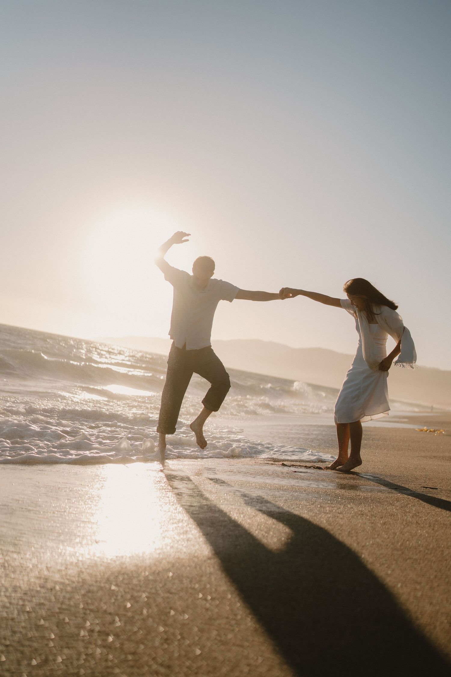 Couple dancing playfully on a sunlit beach at sunset, holding hands with waves and mountains in the background.