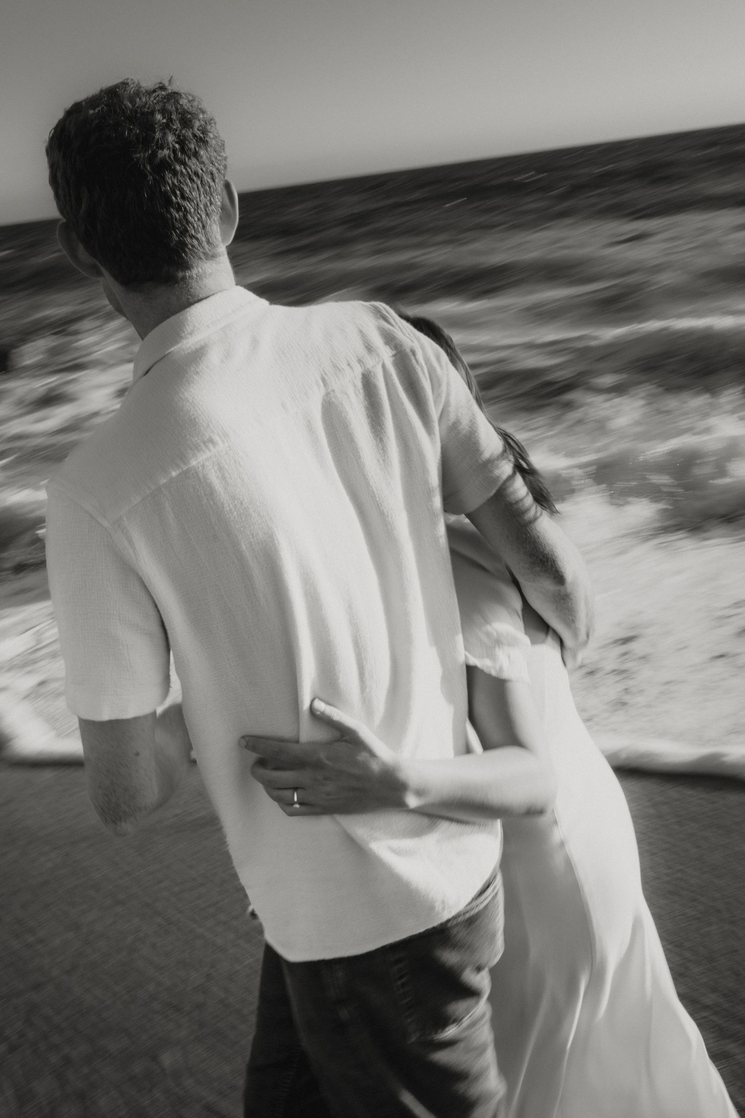 Black and white photo of couple embracing while walking toward ocean waves on sandy beach.