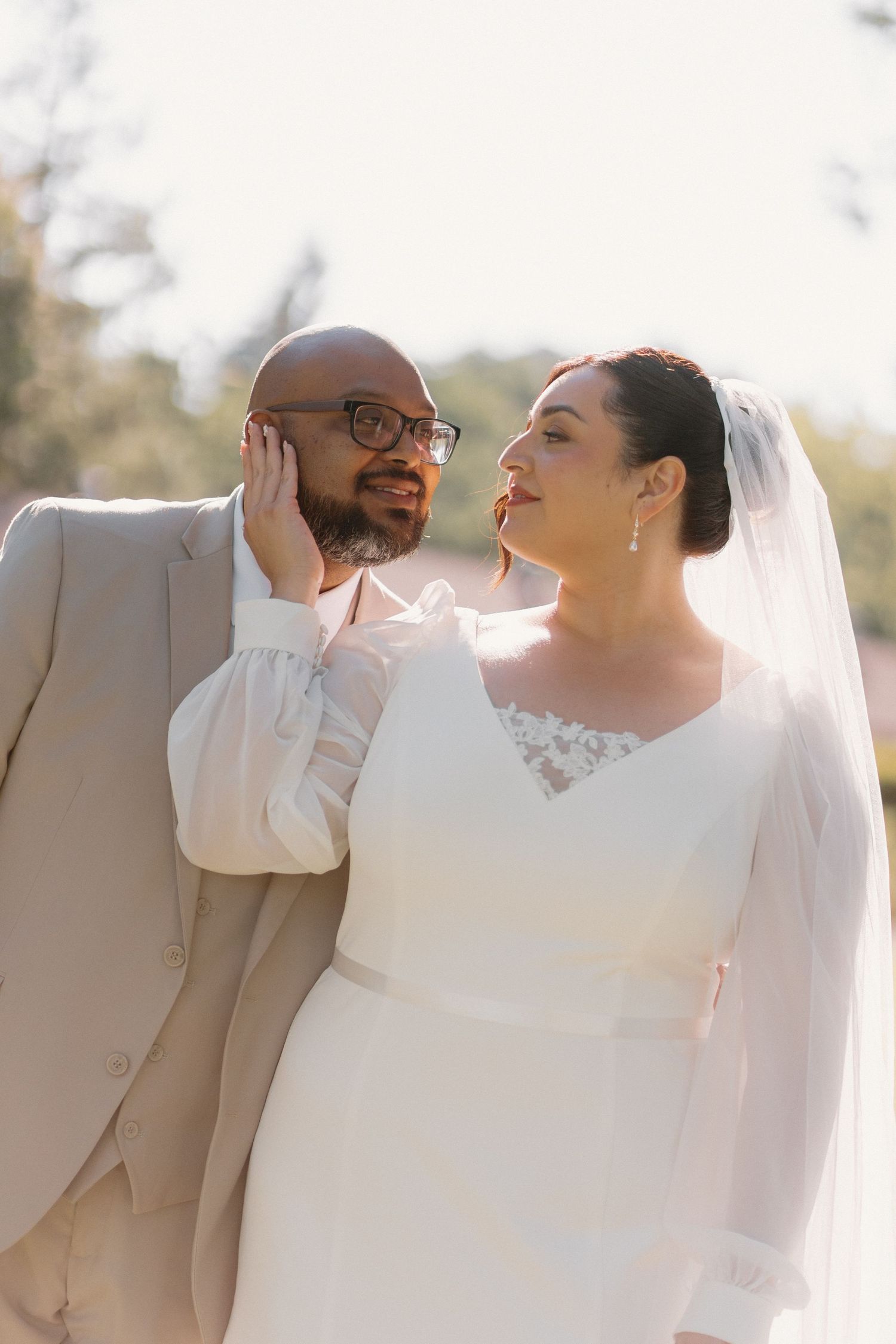 Groom in tan suit and bride in white lace veil gaze at each other in soft sunlight.