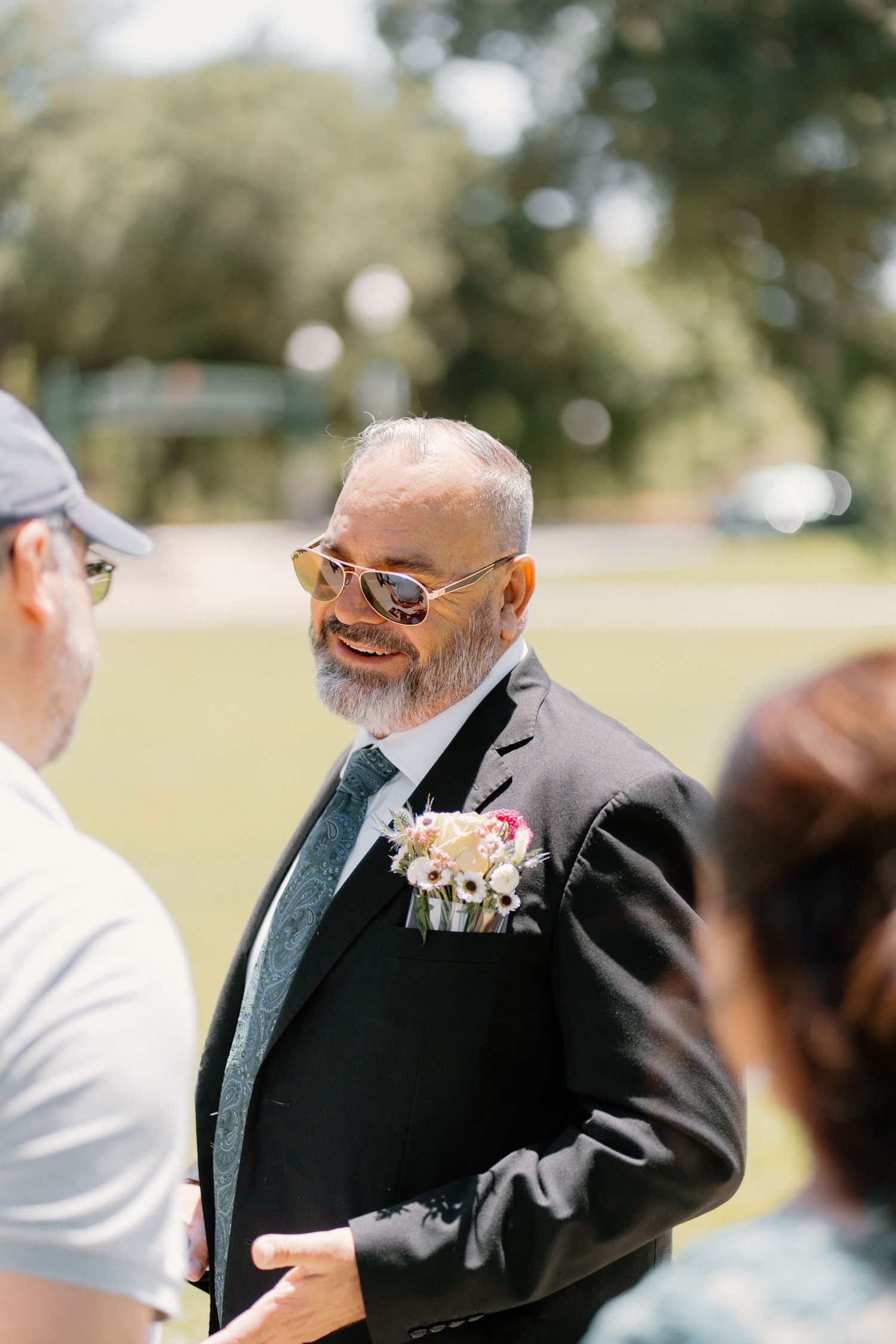 Smiling groom in black suit with floral boutonniere and sunglasses at outdoor wedding ceremony.