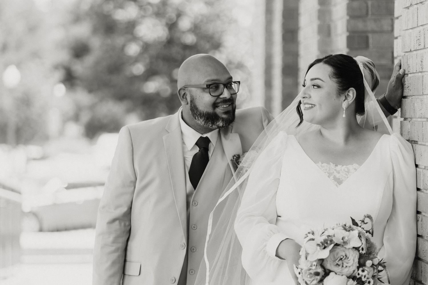 Black and white photo of a smiling bride holding bouquet and groom in suit with glasses at outdoor wedding.