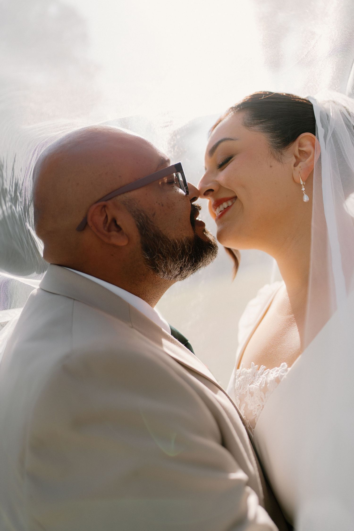 Bride and groom sharing a romantic close moment under veil in soft natural sunlight outdoors.