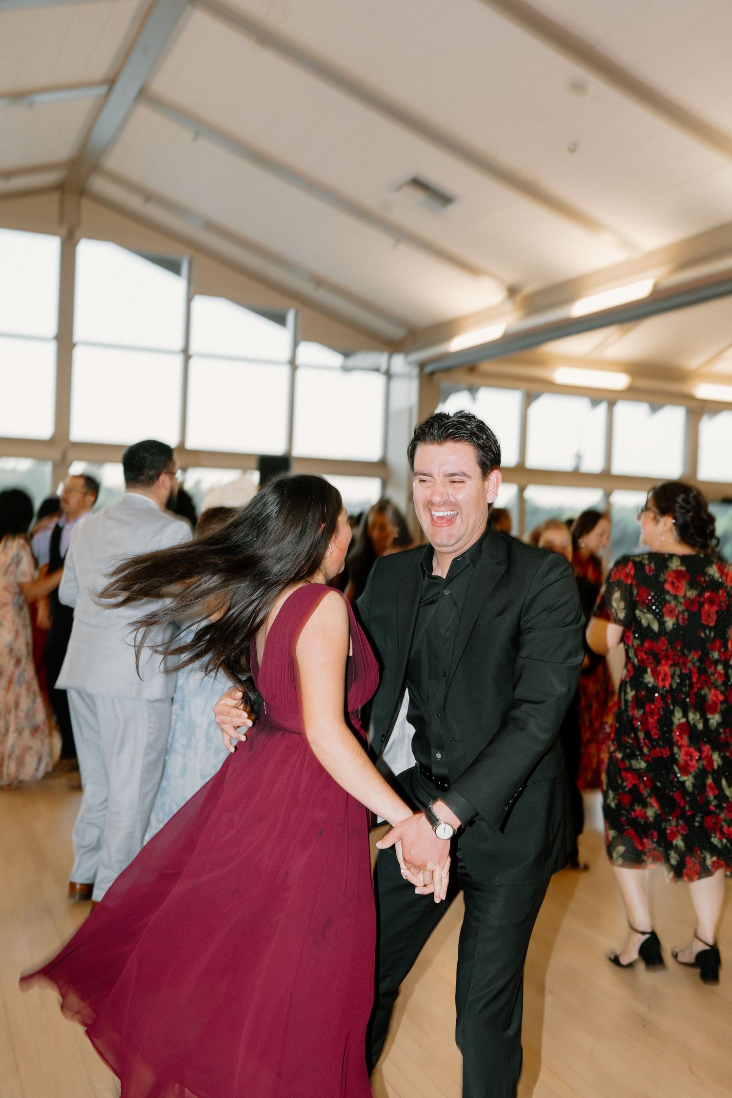 Energetic wedding guest in black suit dancing with woman in burgundy dress at lively reception.