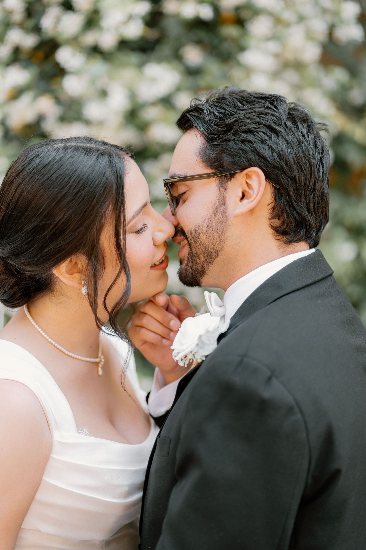 Bride and groom share an intimate moment nose-to-nose in front of lush white floral greenery backdrop.