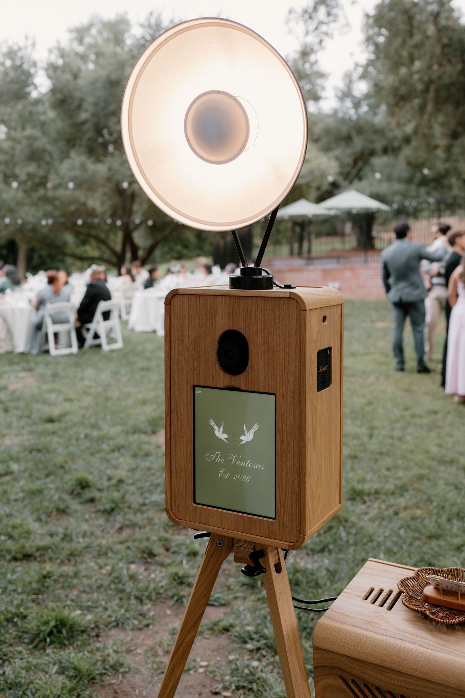 Vintage wooden photo booth with large flash lamp on tripod set up at an outdoor wedding reception.