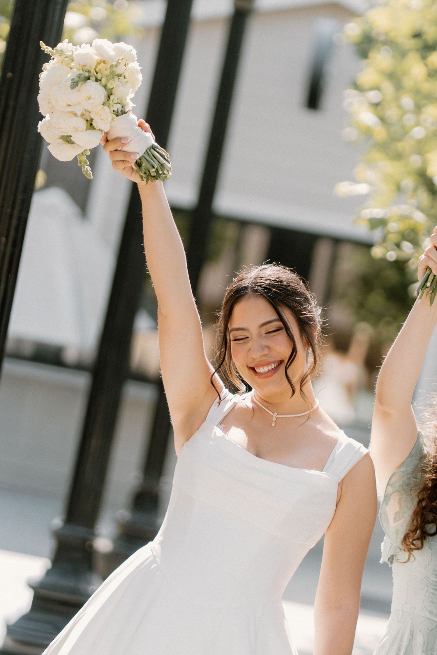Joyful bride in white gown raises bouquet of white flowers outdoors on sunny wedding day.