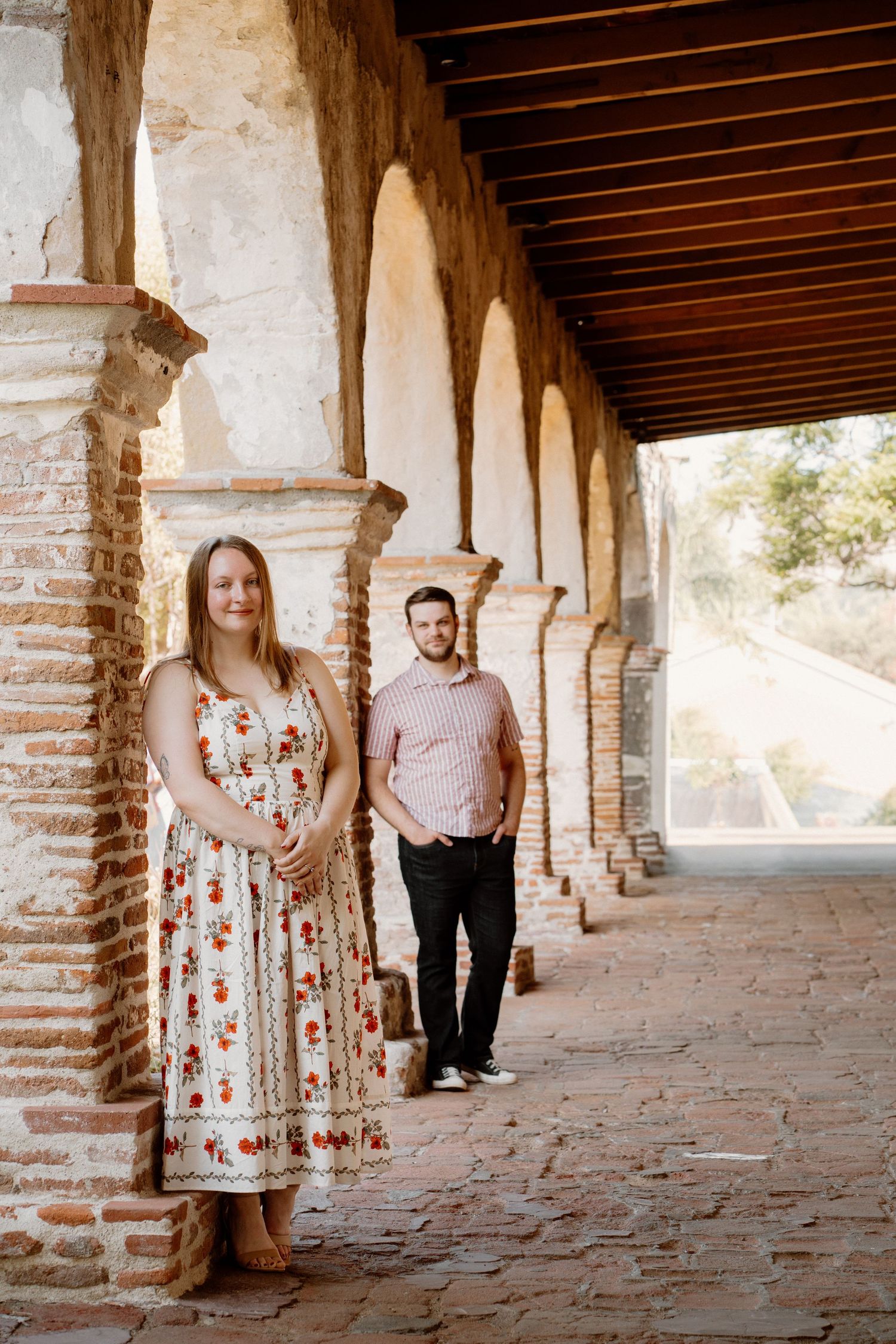 Couple posing under historic brick archway colonnade, woman in floral dress, man in pink shirt.