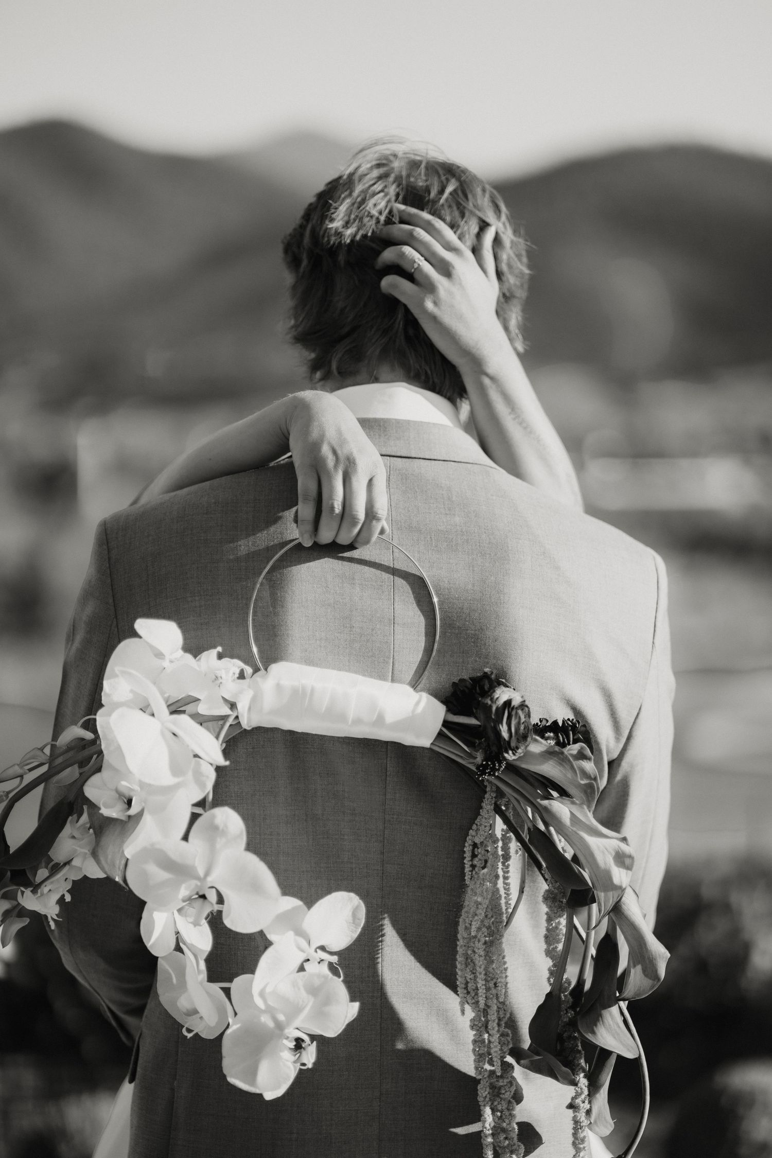 Black and white photo of bride and groom embracing, bride holding floral bouquet with mountains in background.