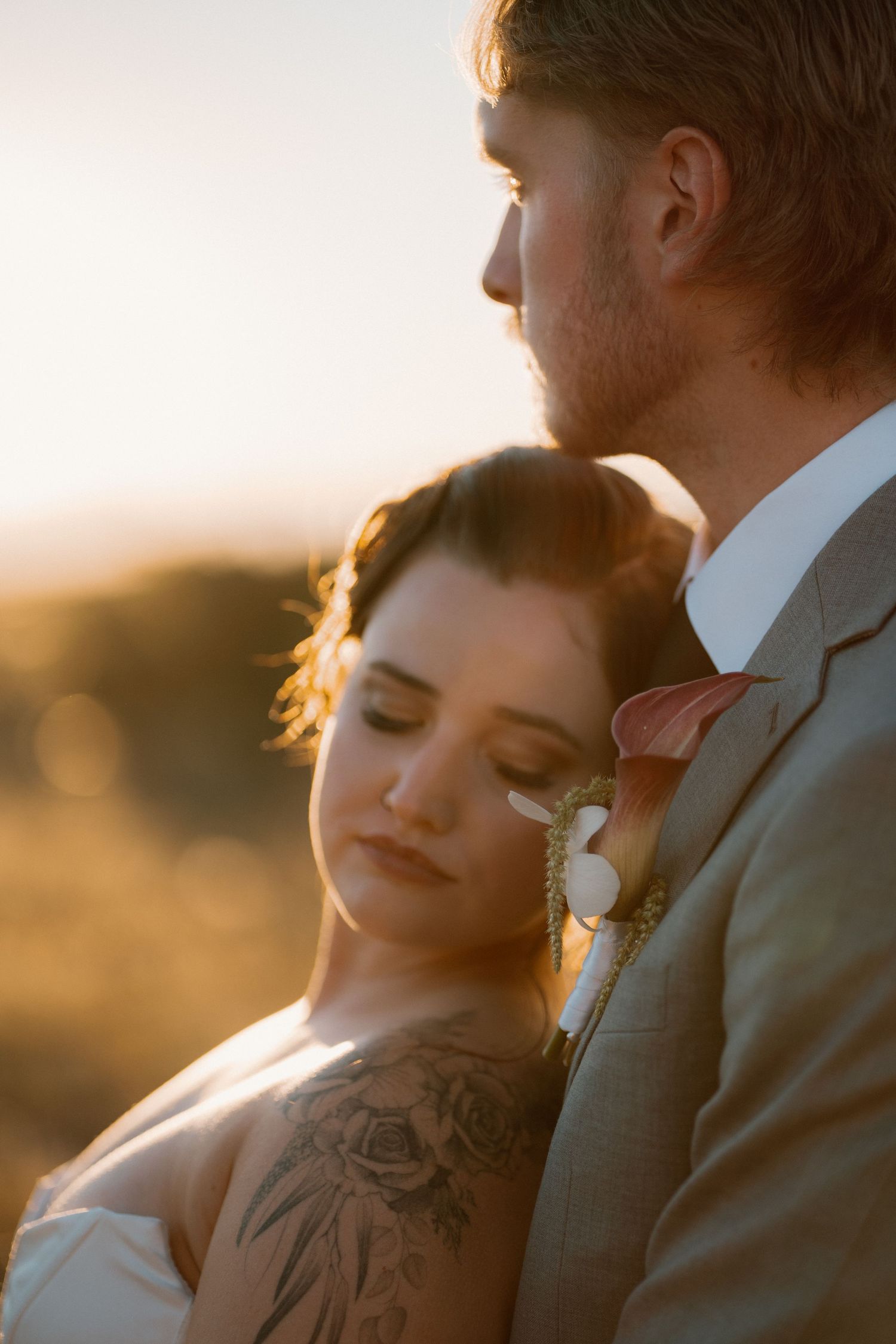 Bride with floral arm tattoo leans against groom in gray suit at sunset, eyes closed in a romantic embrace outdoors.