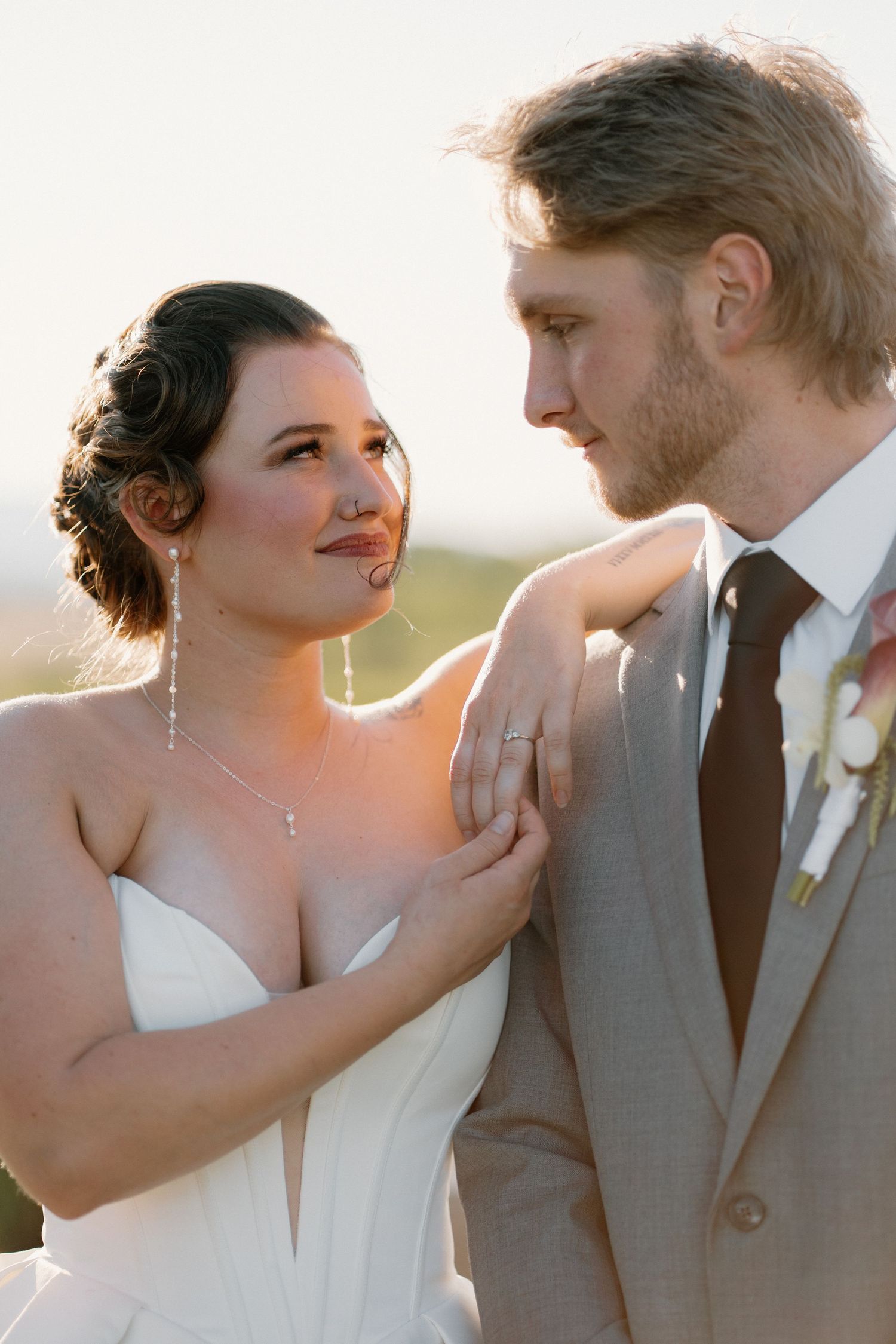 Bride and groom share an intimate moment at golden hour, she wearing a white gown with drop earrings, he in a gray suit.
