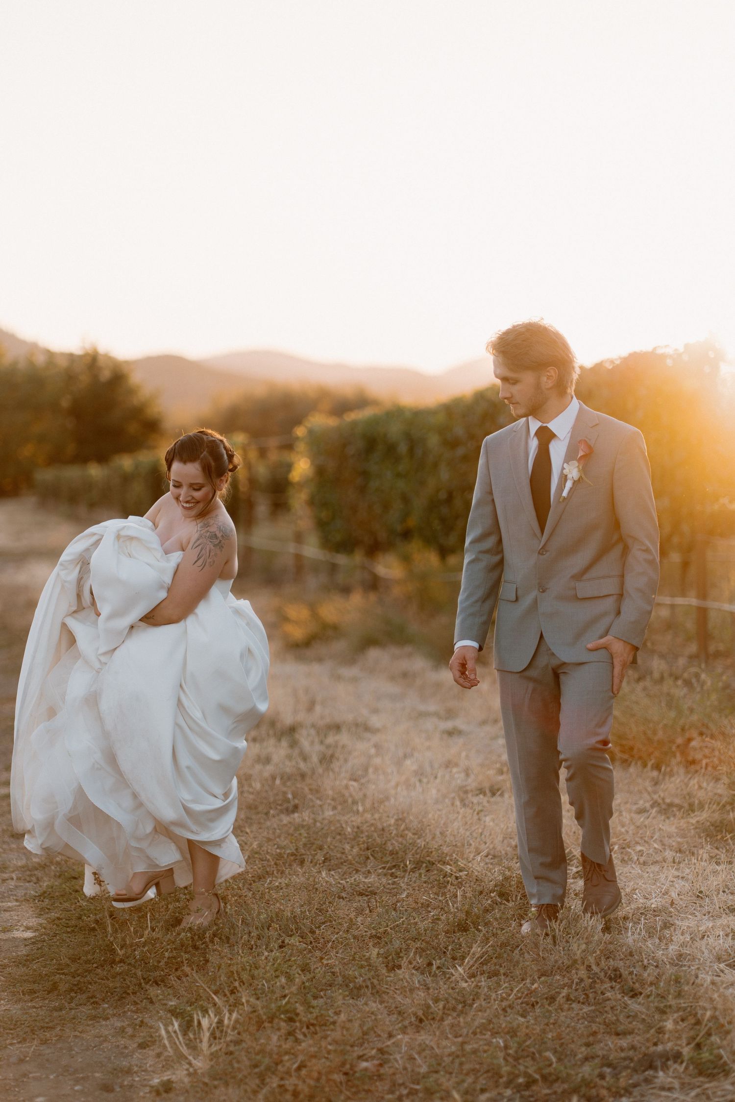 Bride lifts white gown while walking with groom in gray suit through sun-drenched vineyard at golden hour.
