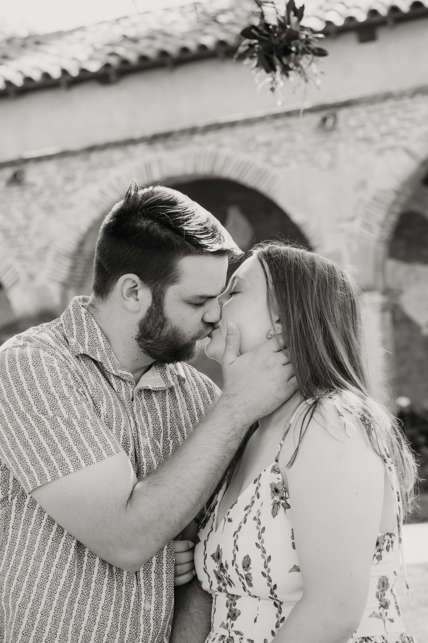 A couple shares a romantic kiss in black and white with a historic stone archway in the background.