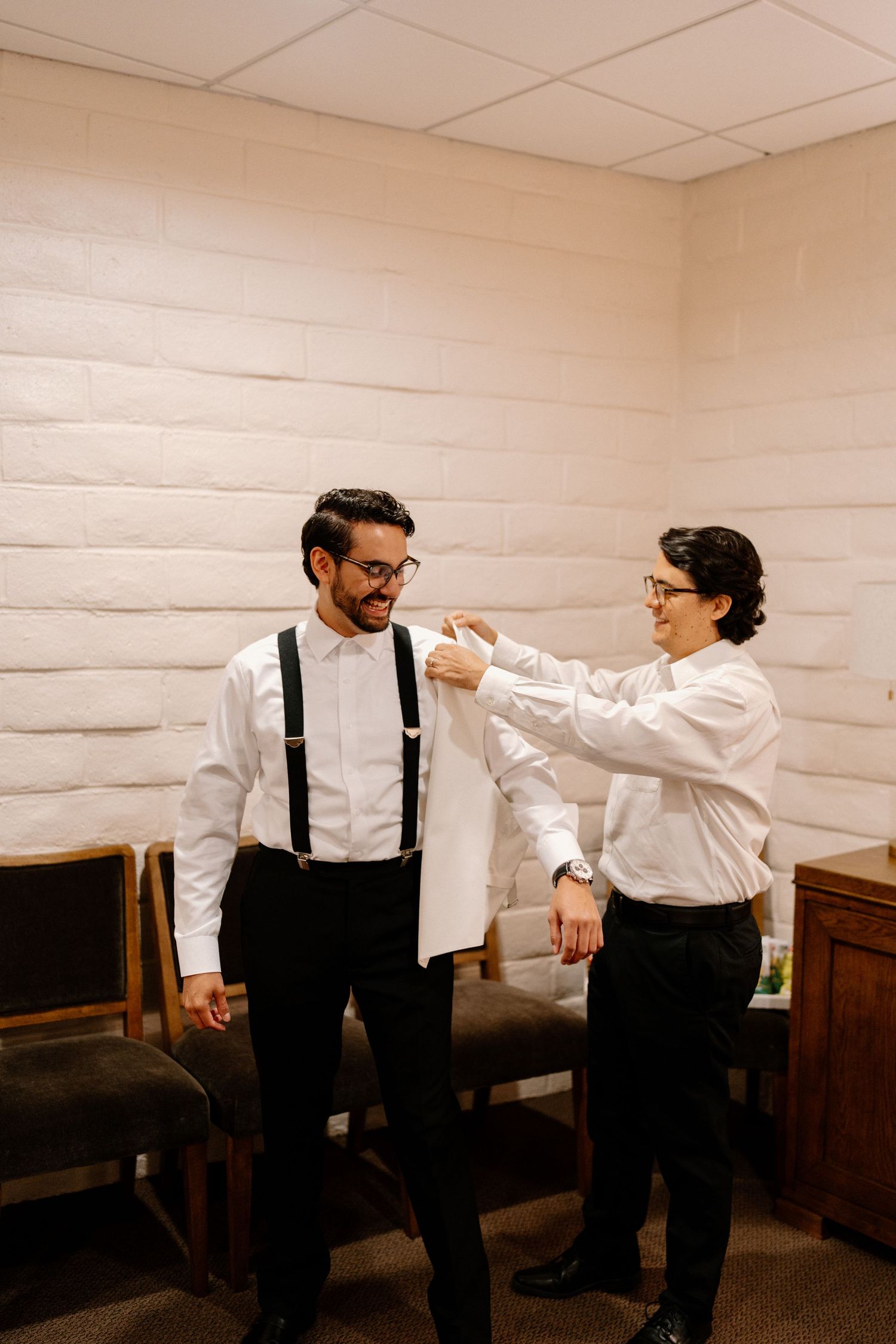 Groomsman helps groom with jacket in a bright room before the wedding ceremony.