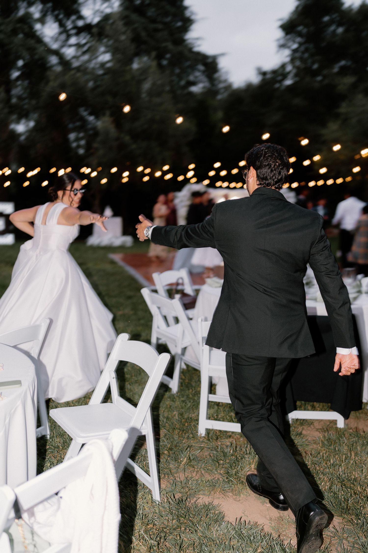 Bride and groom playfully running through white chairs at outdoor evening wedding reception.