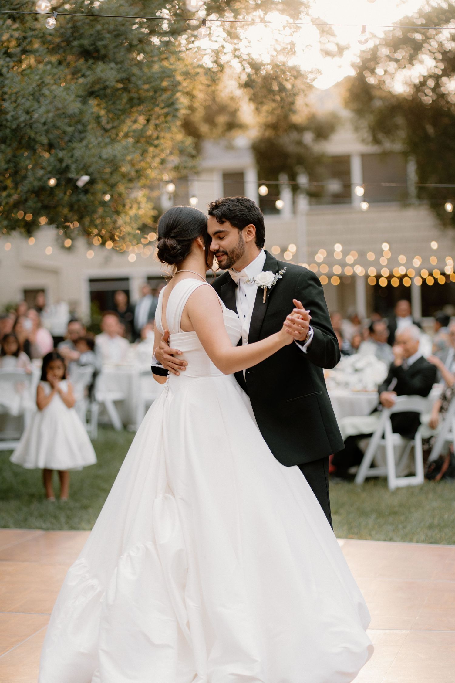 Bride and groom share their first dance at an outdoor wedding reception with string lights glowing in the background.