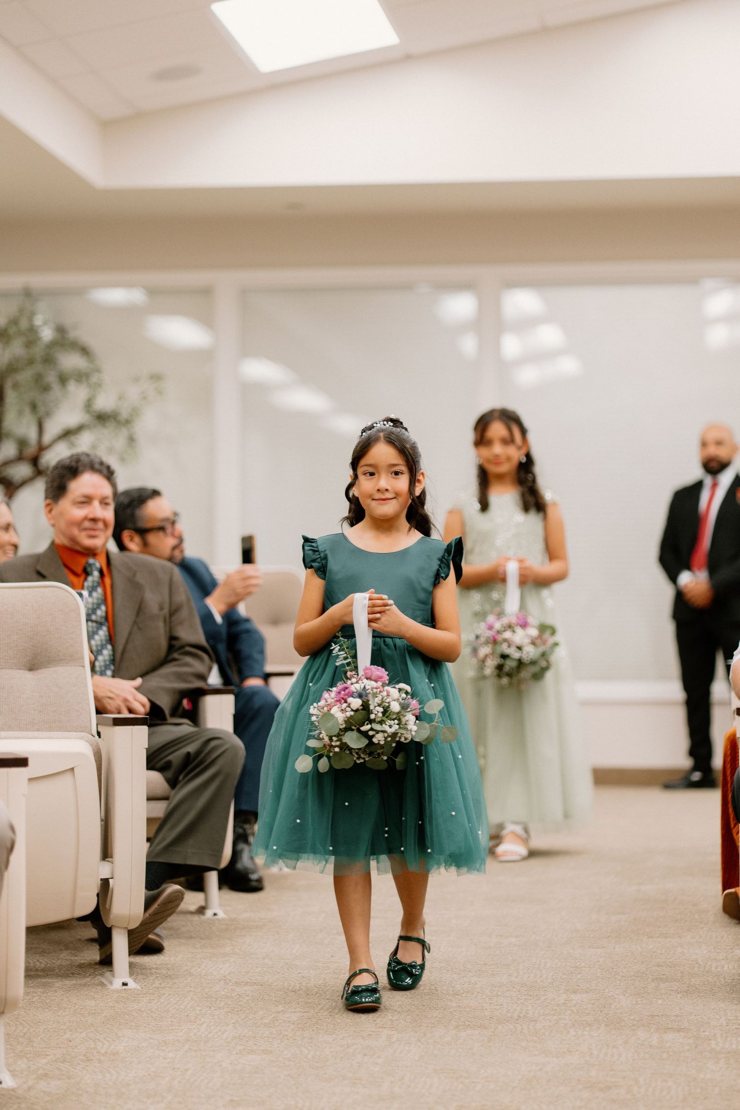 Young flower girl in teal dress walks down wedding aisle carrying floral basket during ceremony.