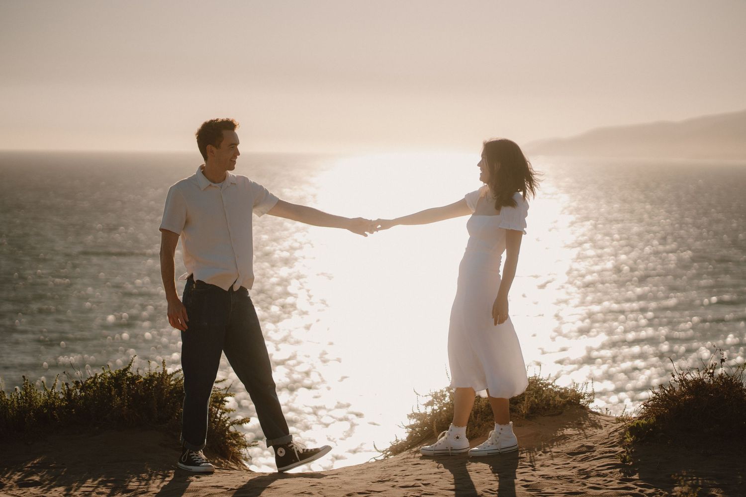 Couple holding hands at sunset on a coastal cliff with sparkling ocean in the background.
