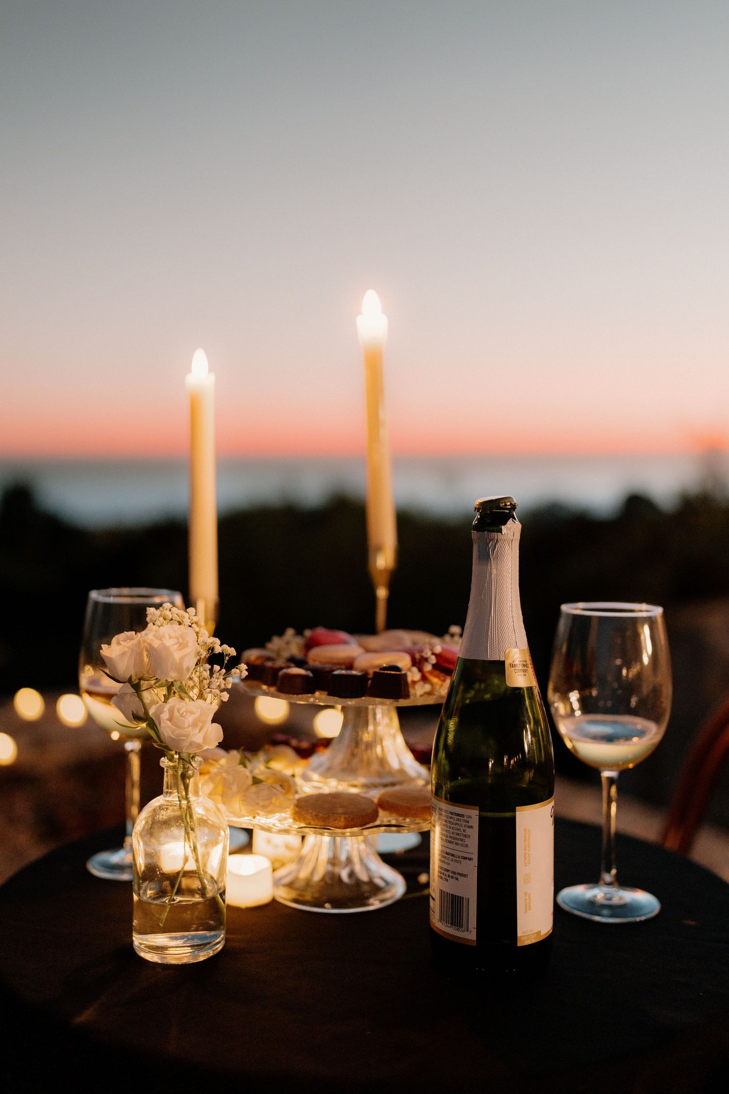 Romantic outdoor proposal setup with champagne, candles, and flowers at sunset by the ocean.
