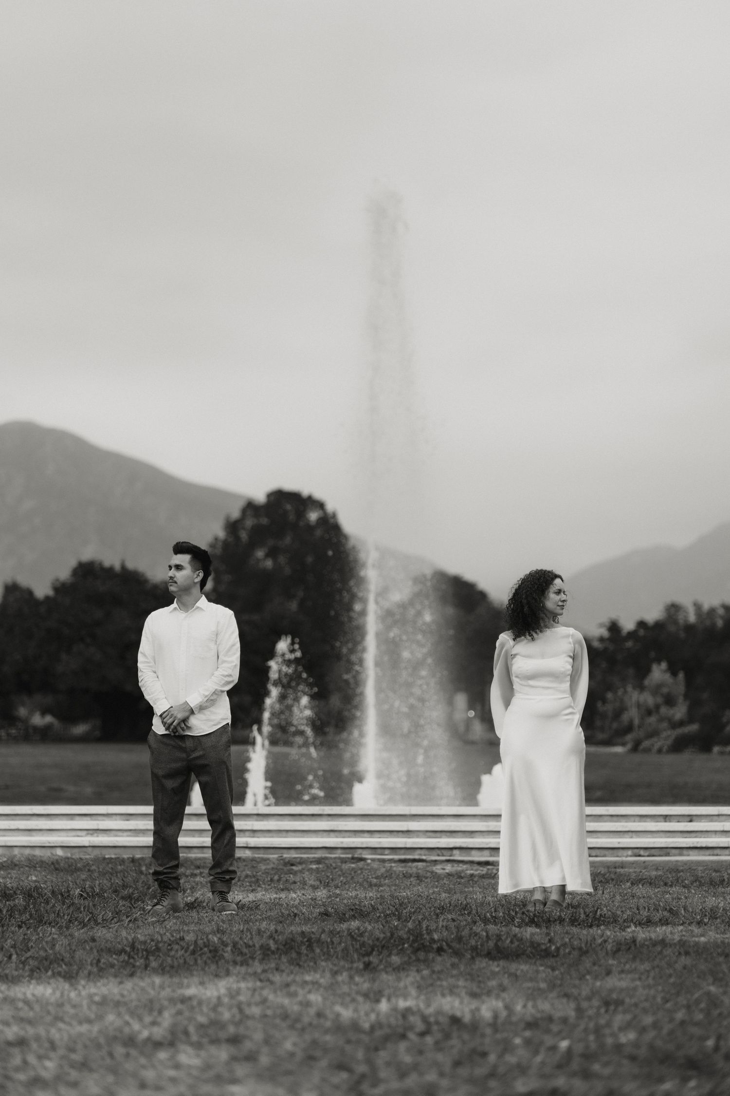 Black and white photo of engaged couple standing apart near a tall fountain with mountains behind.