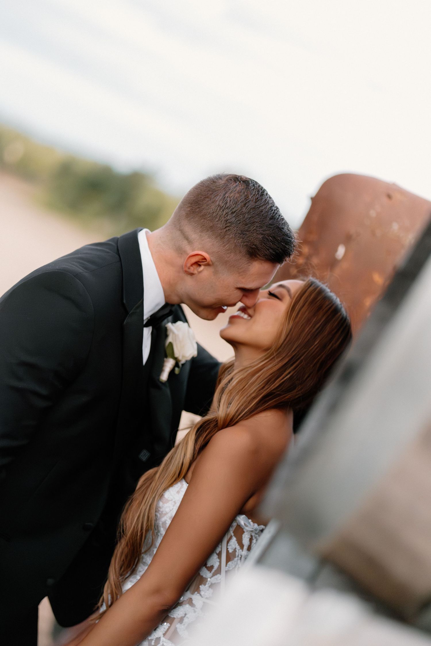 Bride and groom sharing a romantic almost-kiss moment leaning against a rustic wall at golden hour.
