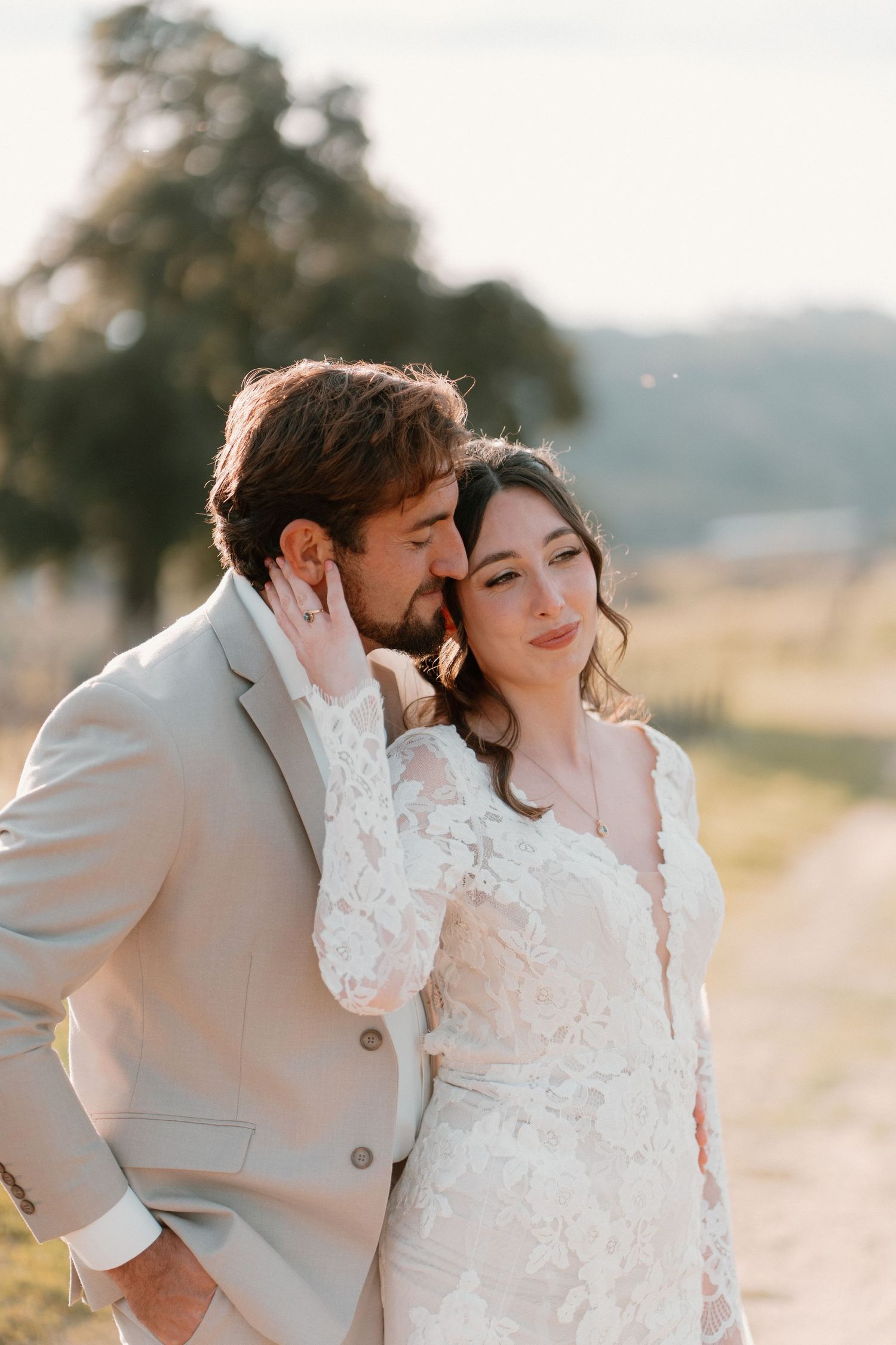 Groom tenderly kisses bride on the cheek as she leans into him during golden hour outdoor wedding portrait.