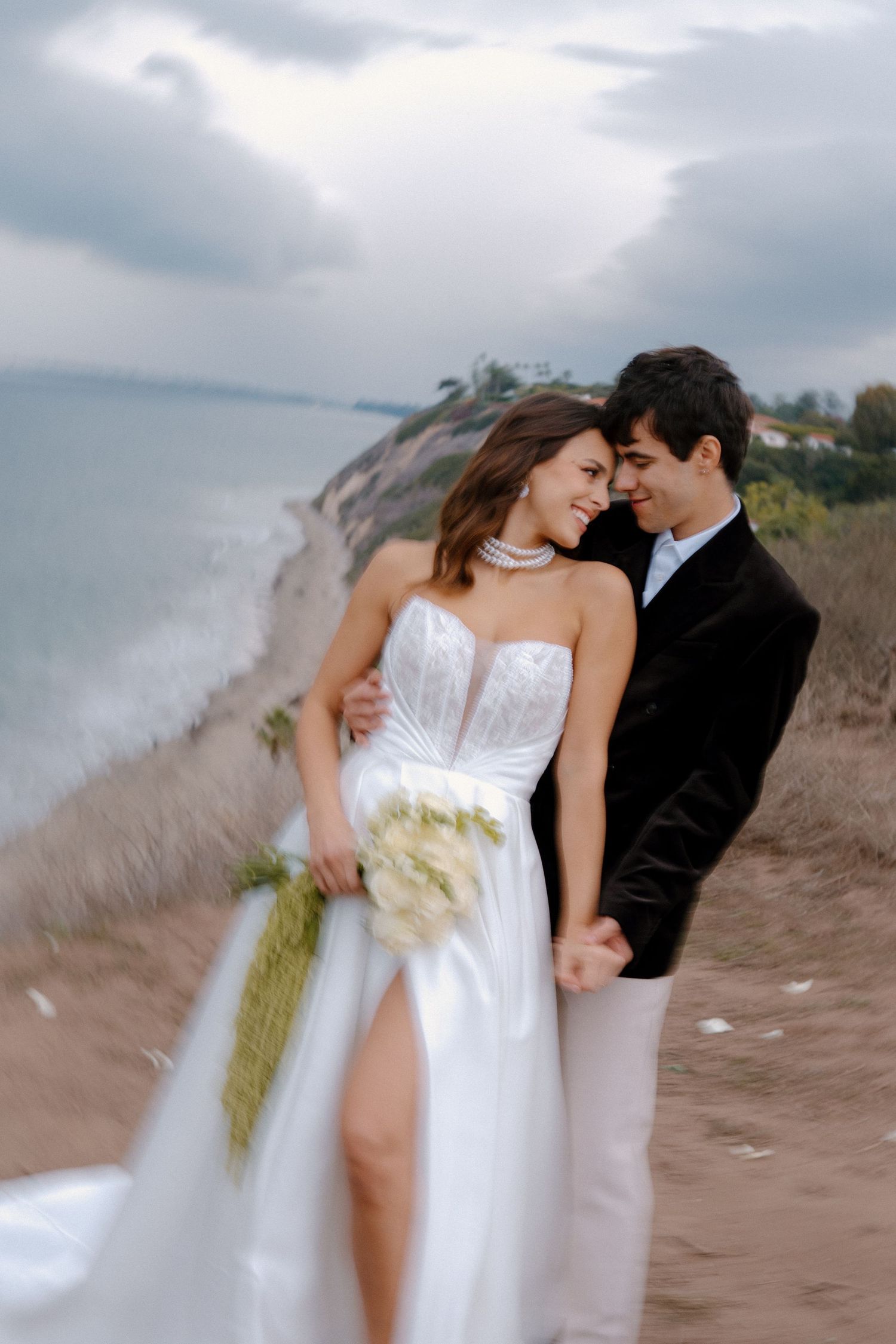 Elegant bride and groom embrace on a coastal cliff, she wears a white strapless gown with slit holding a bouquet.